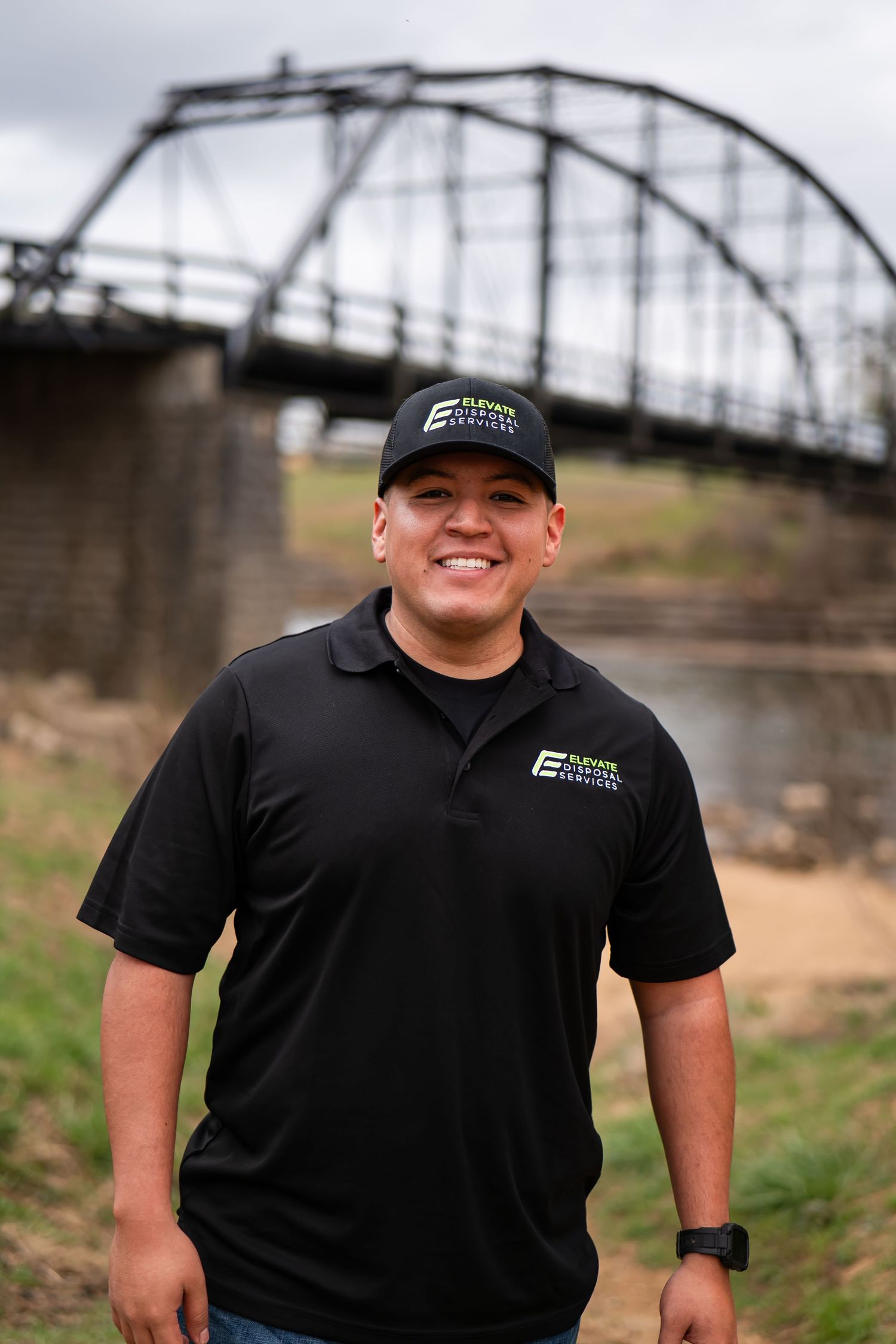 Team member wearing black uniform and baseball cap standing near a metal bridge structure.
