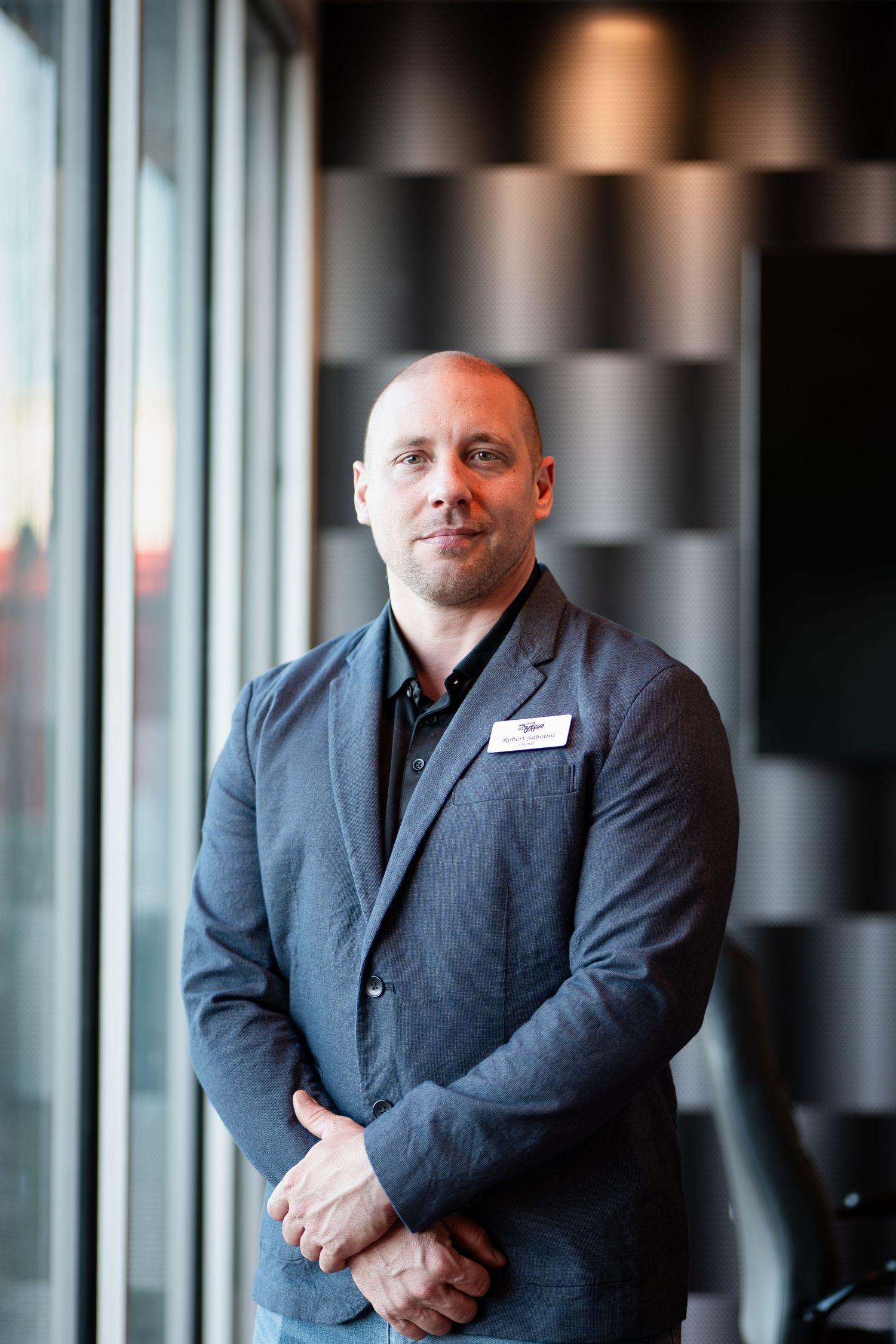 Professional portrait of a person in a gray suit jacket standing by windows.