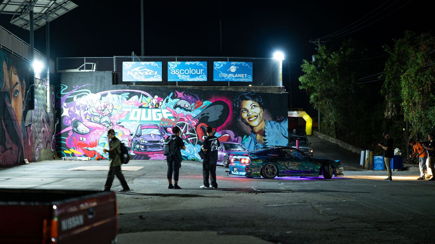 Nighttime street scene with graffiti walls and scattered people walking.