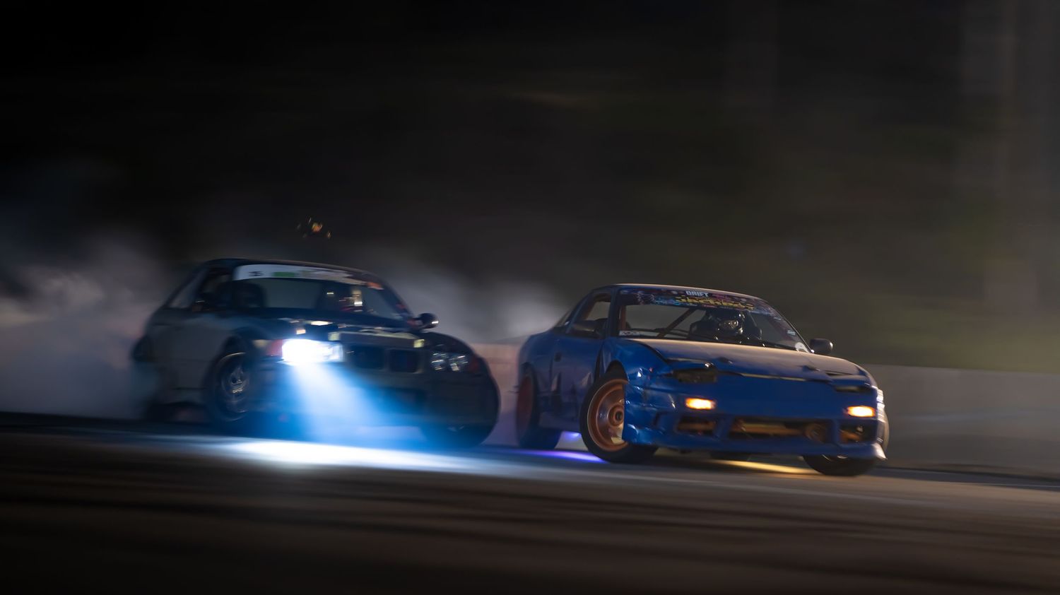 Night time drift racing with cars creating light trails and smoke effects.