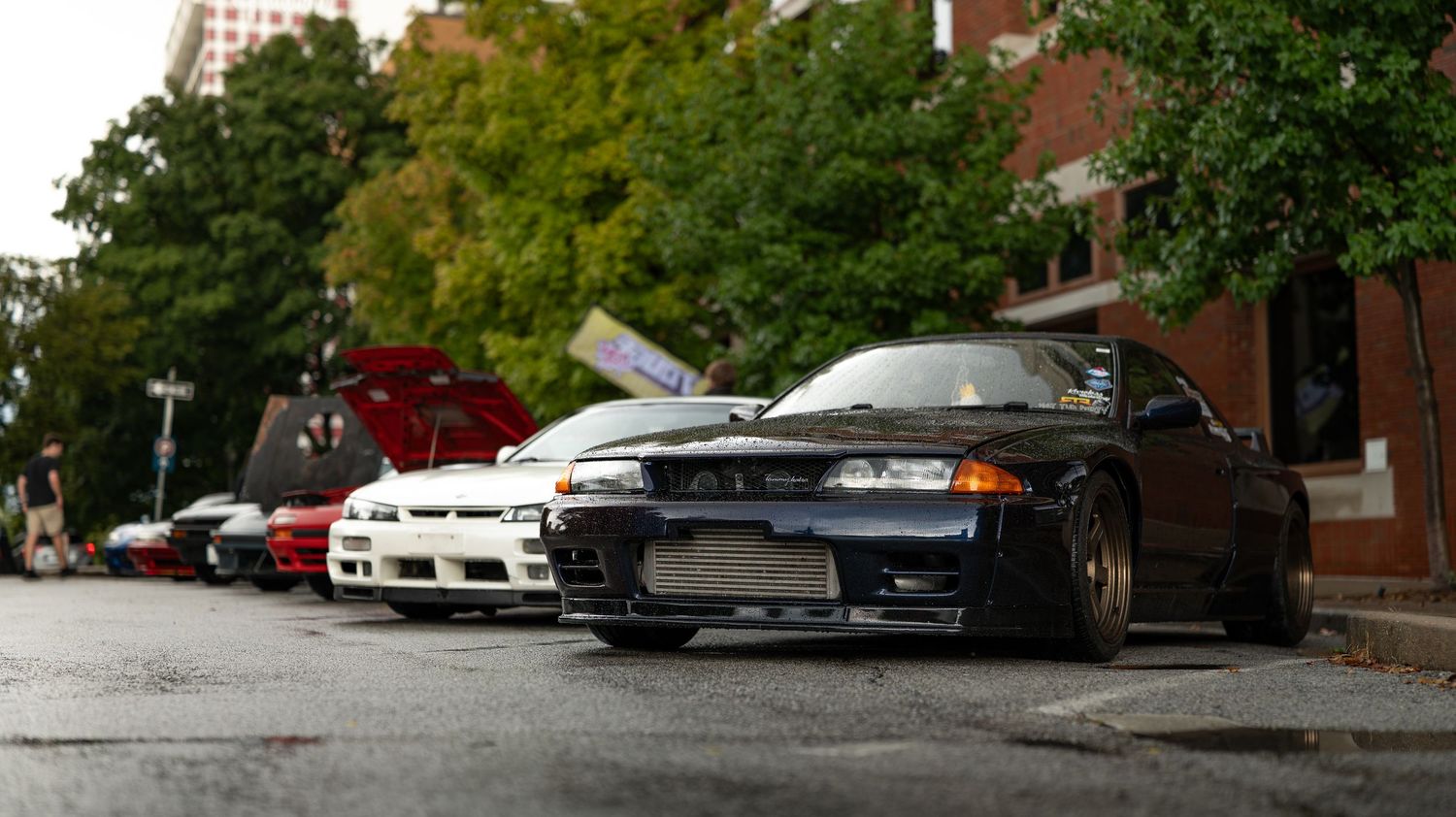 Wide view of cars displayed at a car meet against brick buildings.