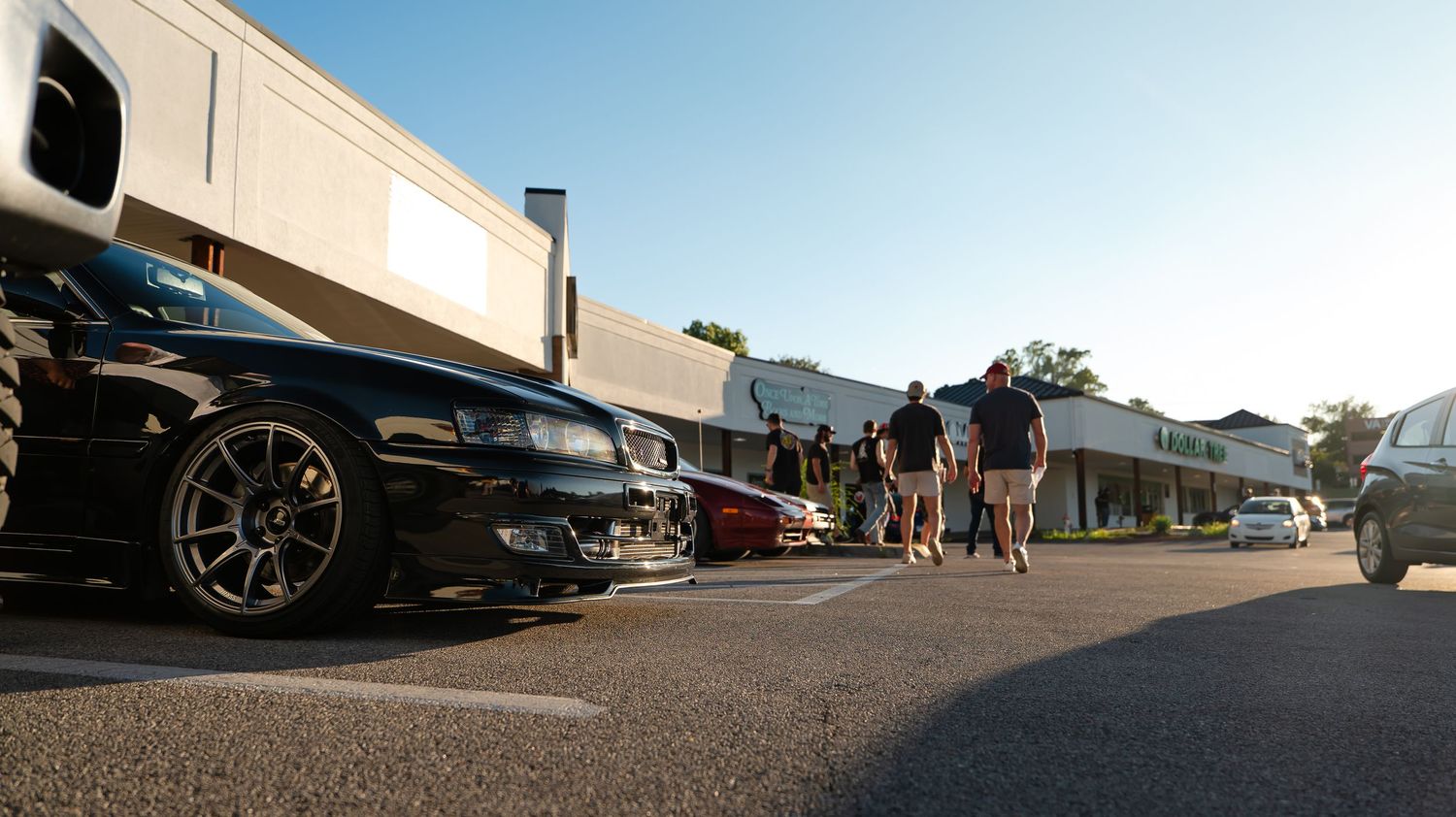 Modified cars parked in a row outside storefronts on a sunny day.