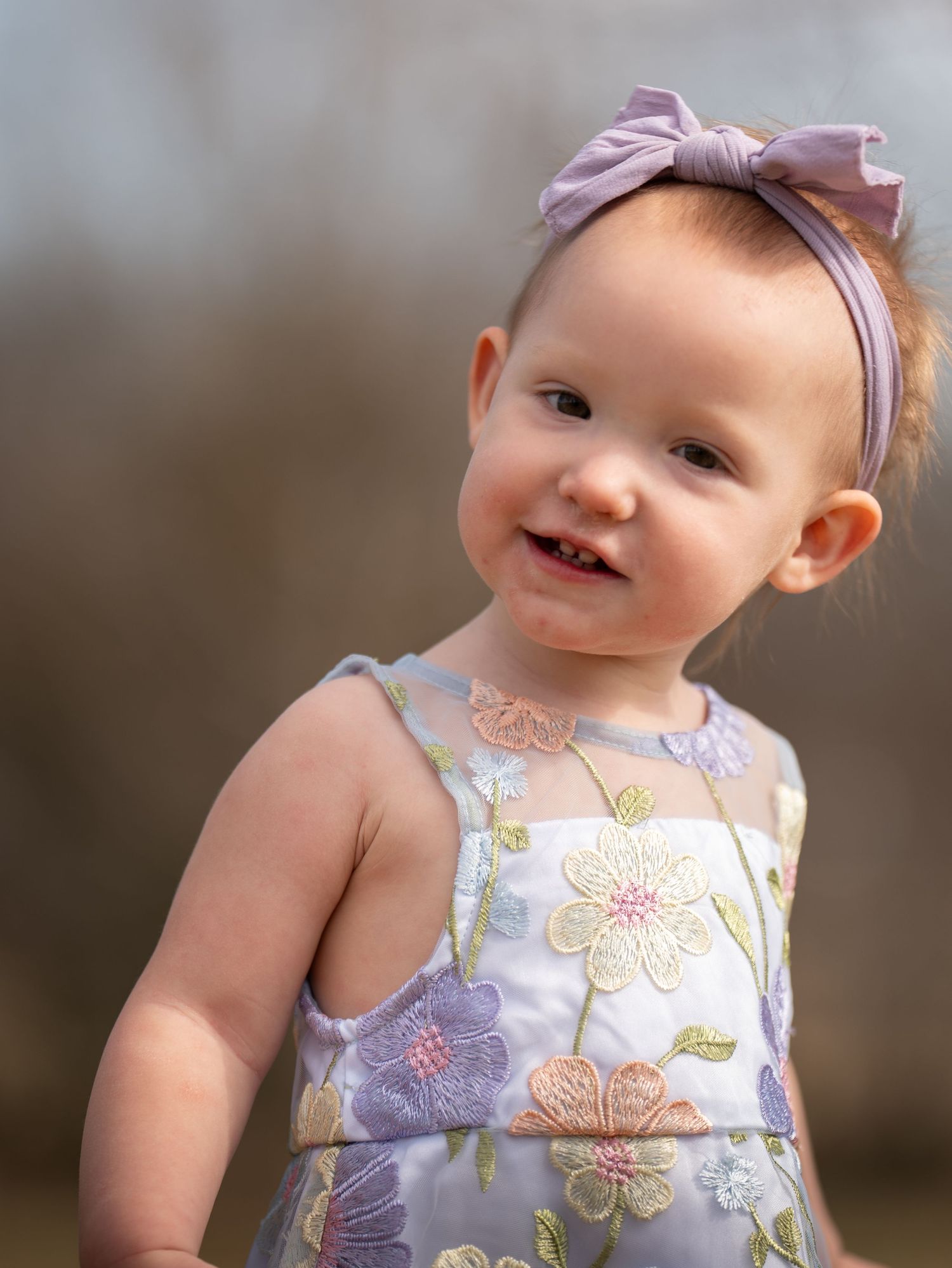 Smiling baby girl in floral dress and purple bow headband outdoors.