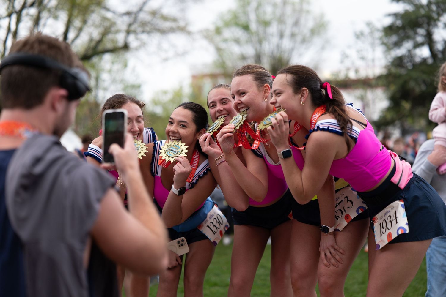 Women in pink running gear pose with medals after a race while a man photographs them outdoors.