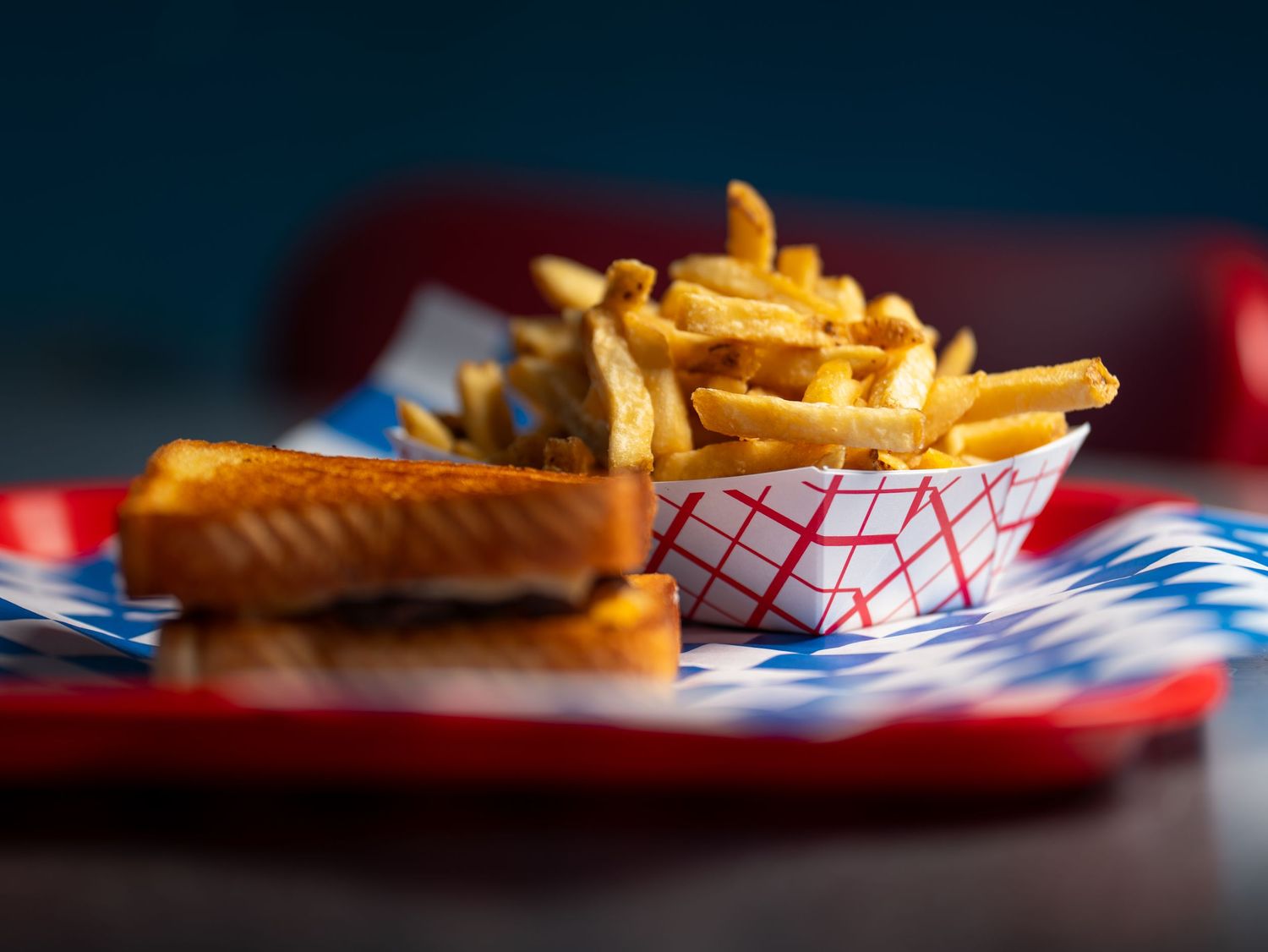 A close-up shot of crispy french fries and sandwich in a red paper tray against a dark background.