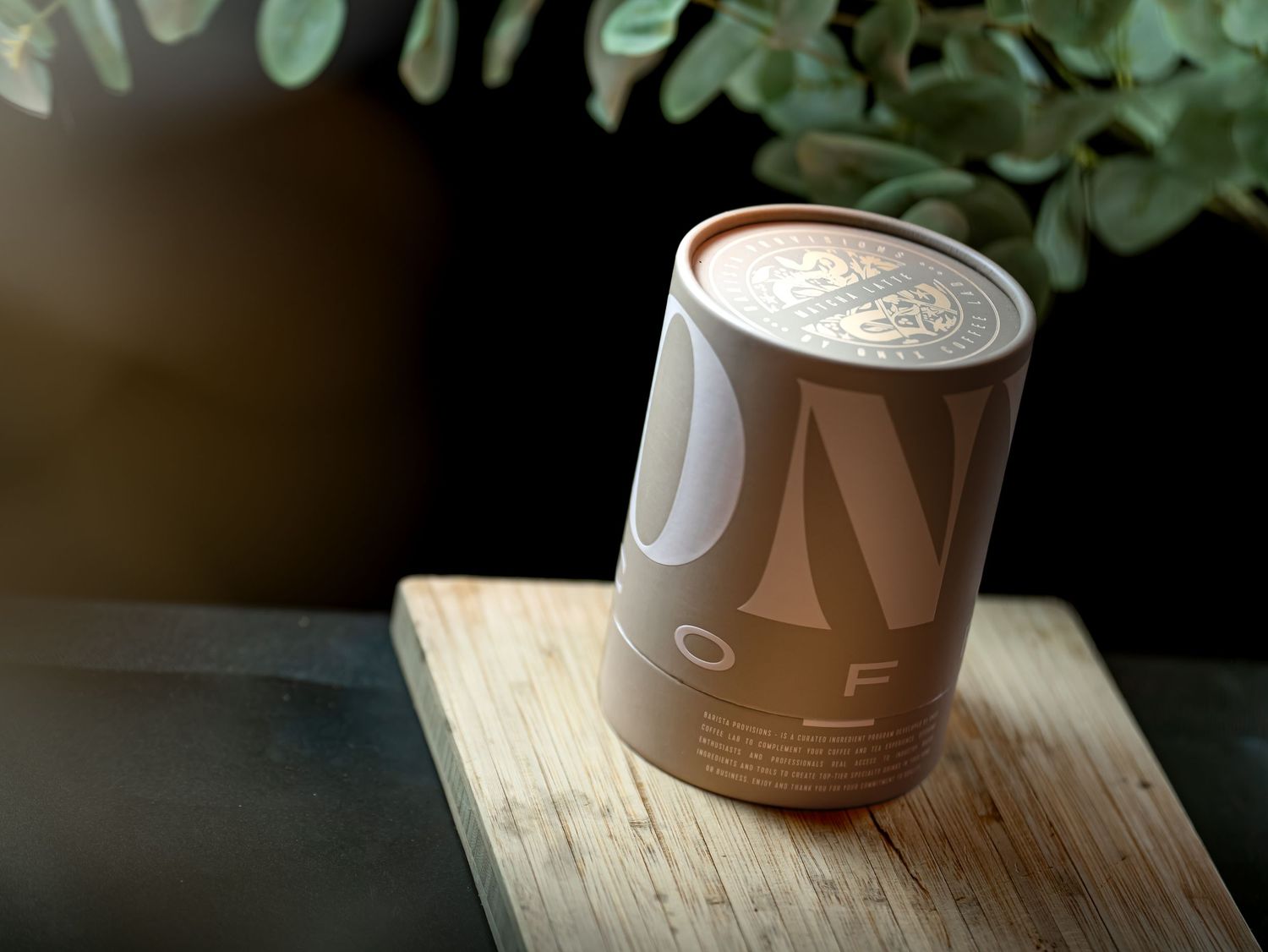 A cylindrical brown coffee canister with elegant typography sits on a wooden board beside lush green foliage.