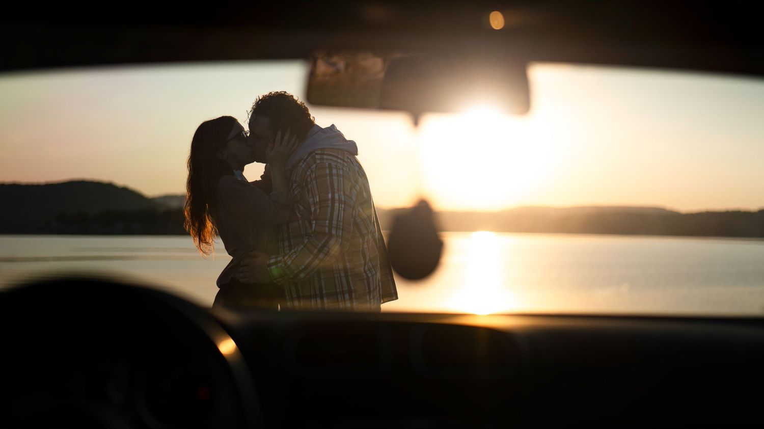 Silhouetted figures watching sunset through car windshield near water.