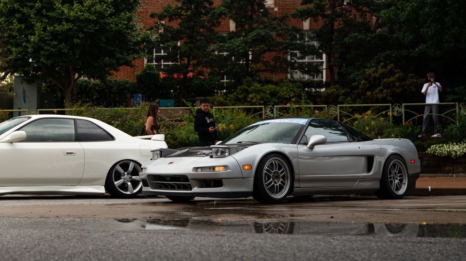 White sedan and silver NSX sports car parked on wet pavement.