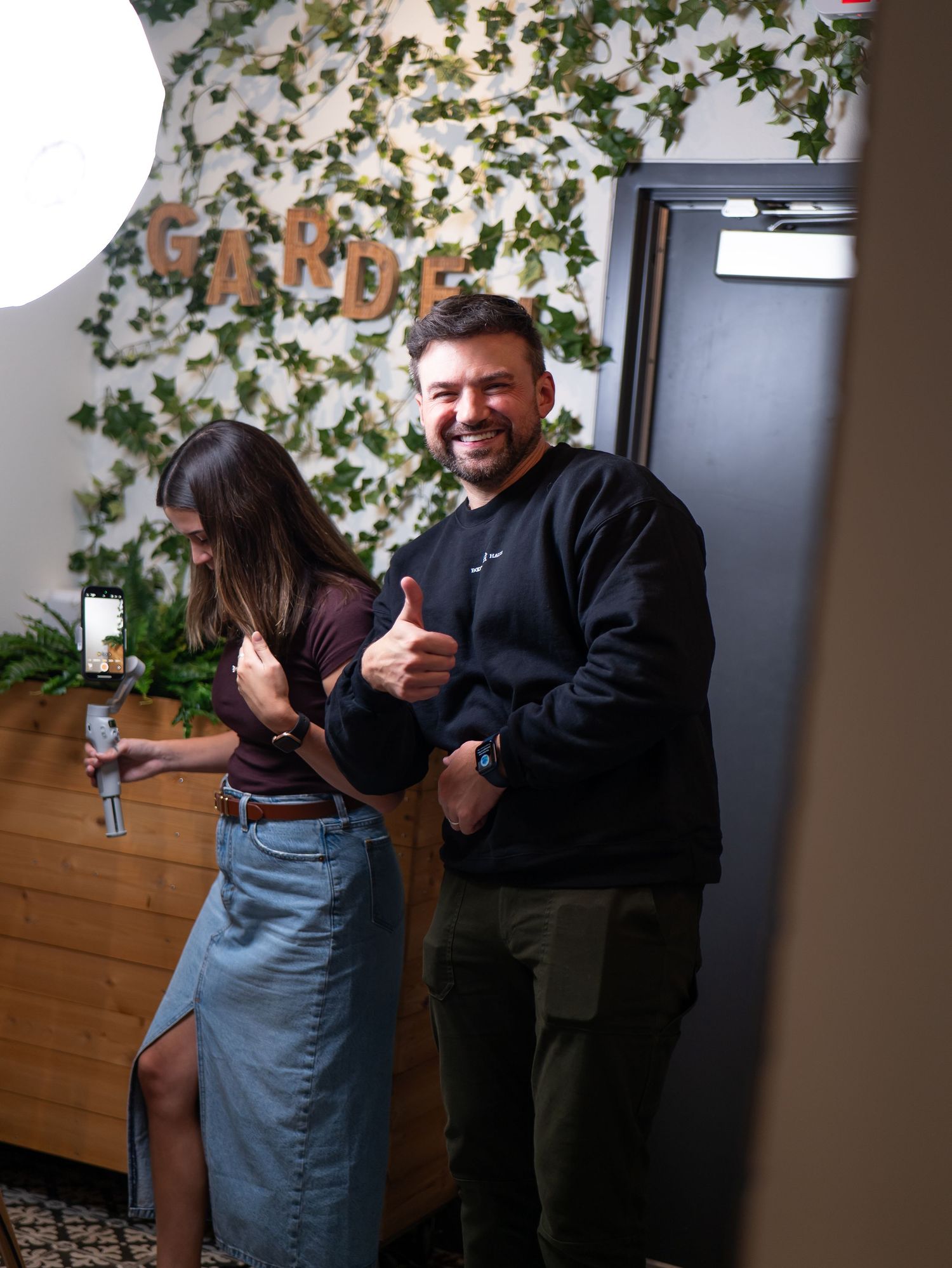 Two people in a casual indoor setting with wooden paneling and greenery decorations sharing a playful moment together.