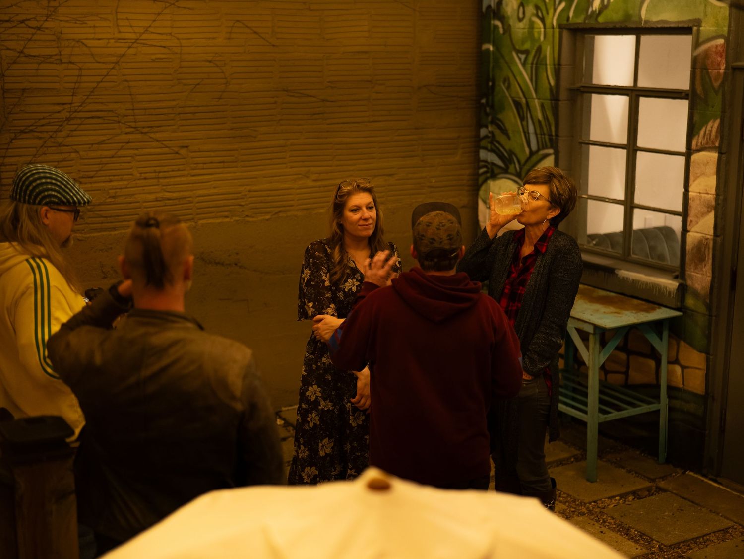 A group of people gather in a dimly lit room with wooden walls and a window, creating a warm atmospheric scene.
