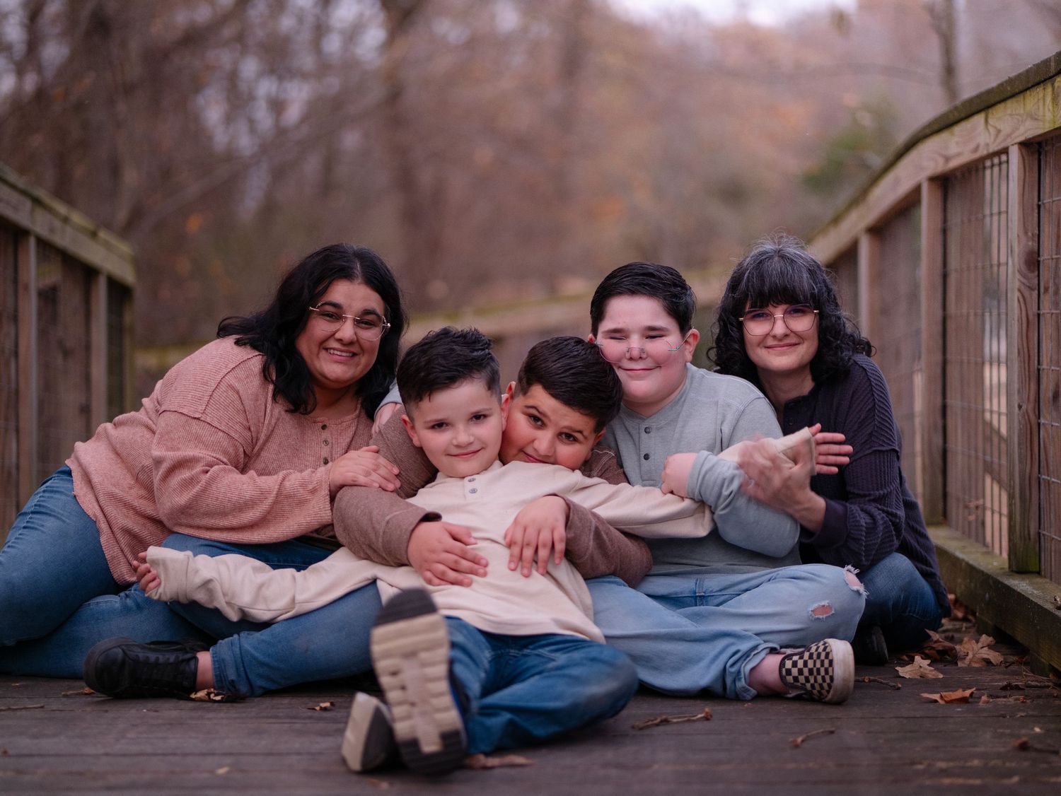 A family lies down together on a wooden bridge in an autumn forest setting for a casual outdoor photo session.