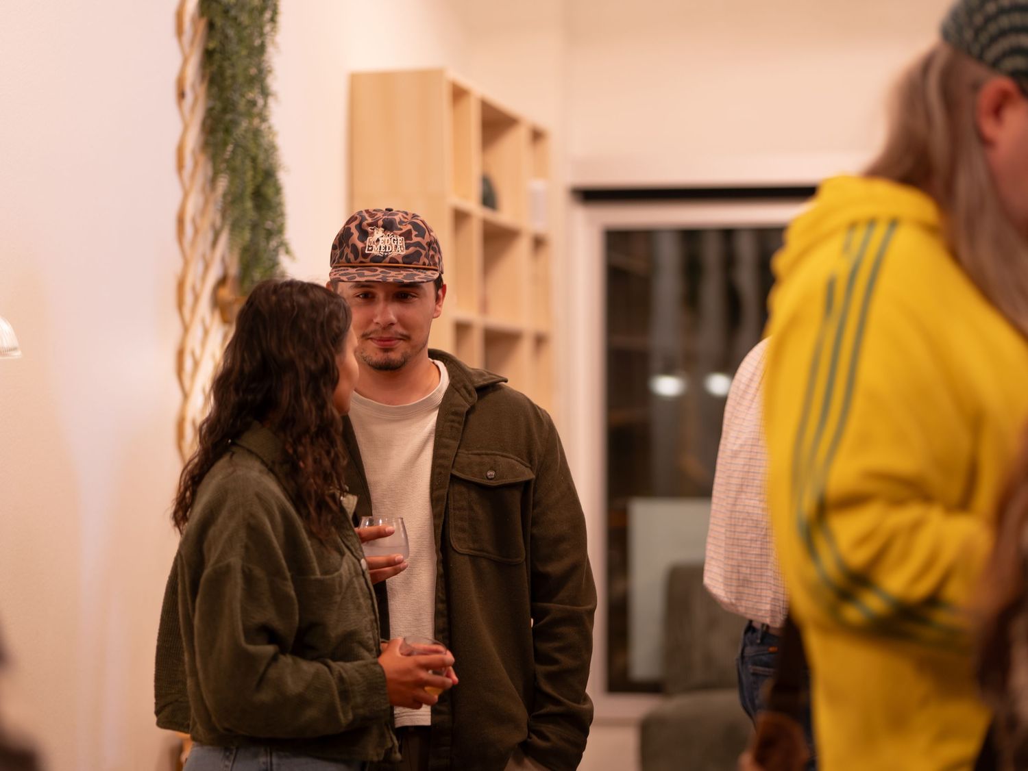 People gather and socialize at an indoor event with white walls and yellow accents in the background.