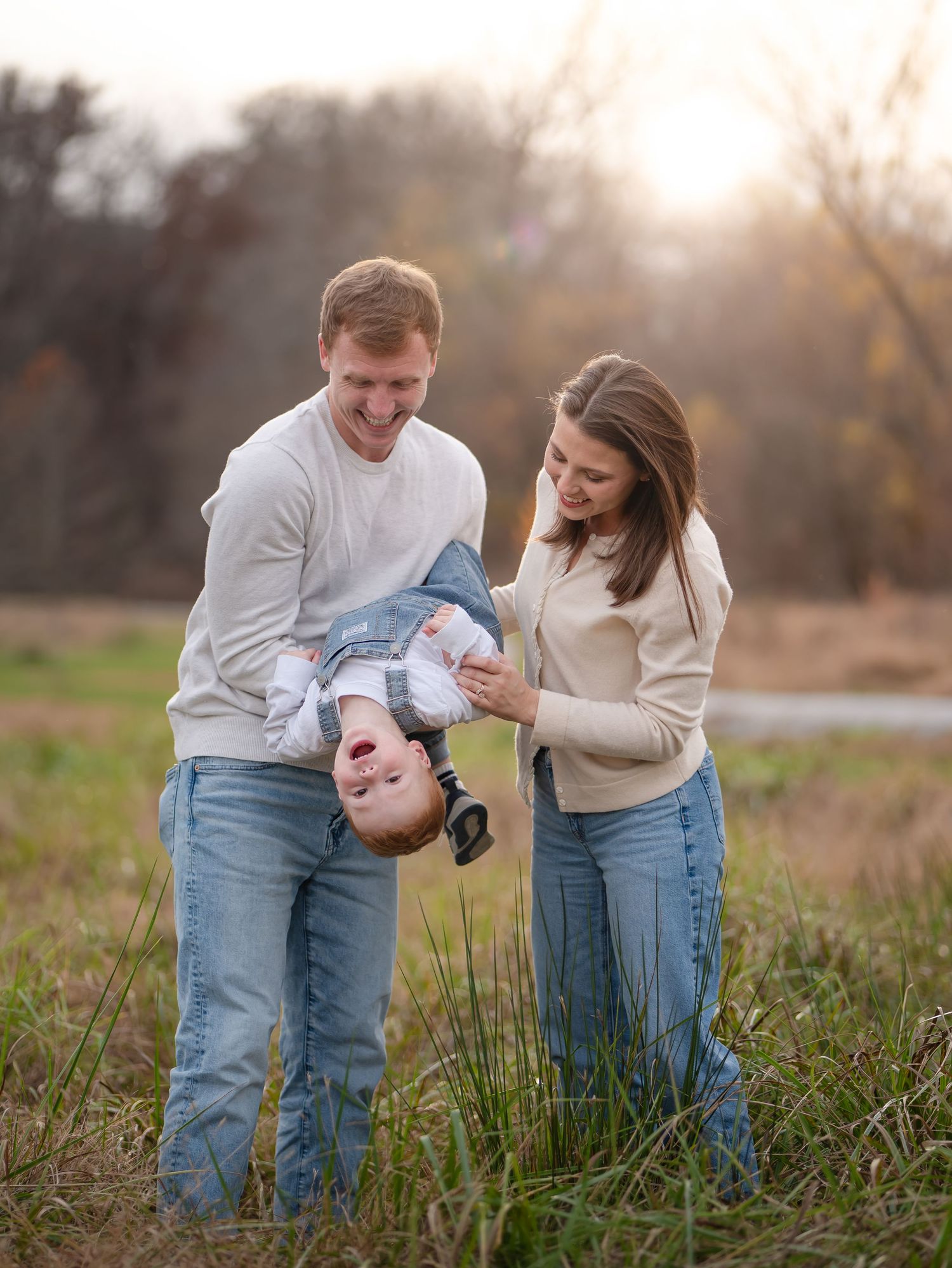 A couple in matching white sweaters and jeans share tender moments with their baby in a grassy field at sunset.