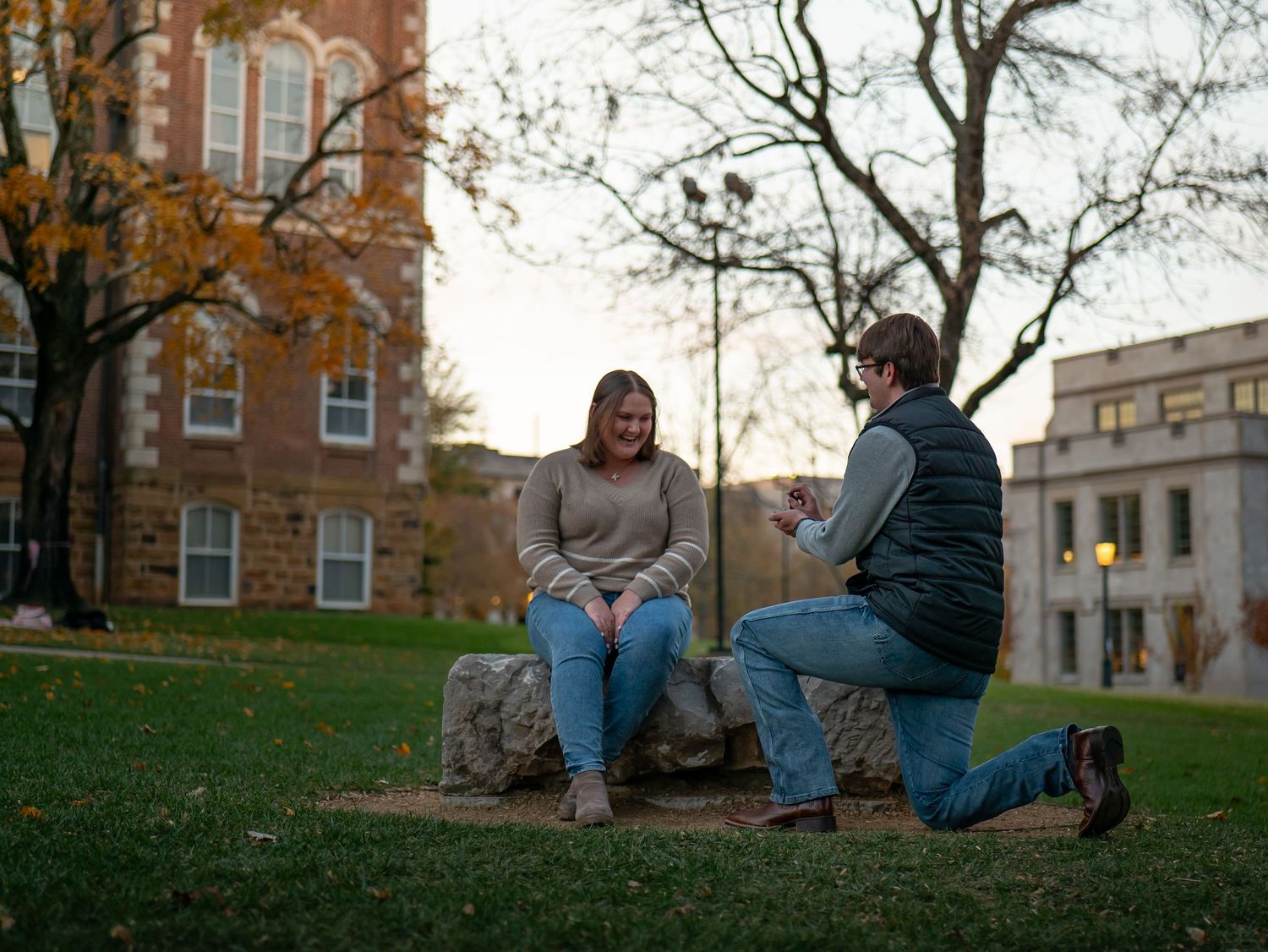 Two people sit together on a grassy hill during autumn with brick buildings visible in the background.