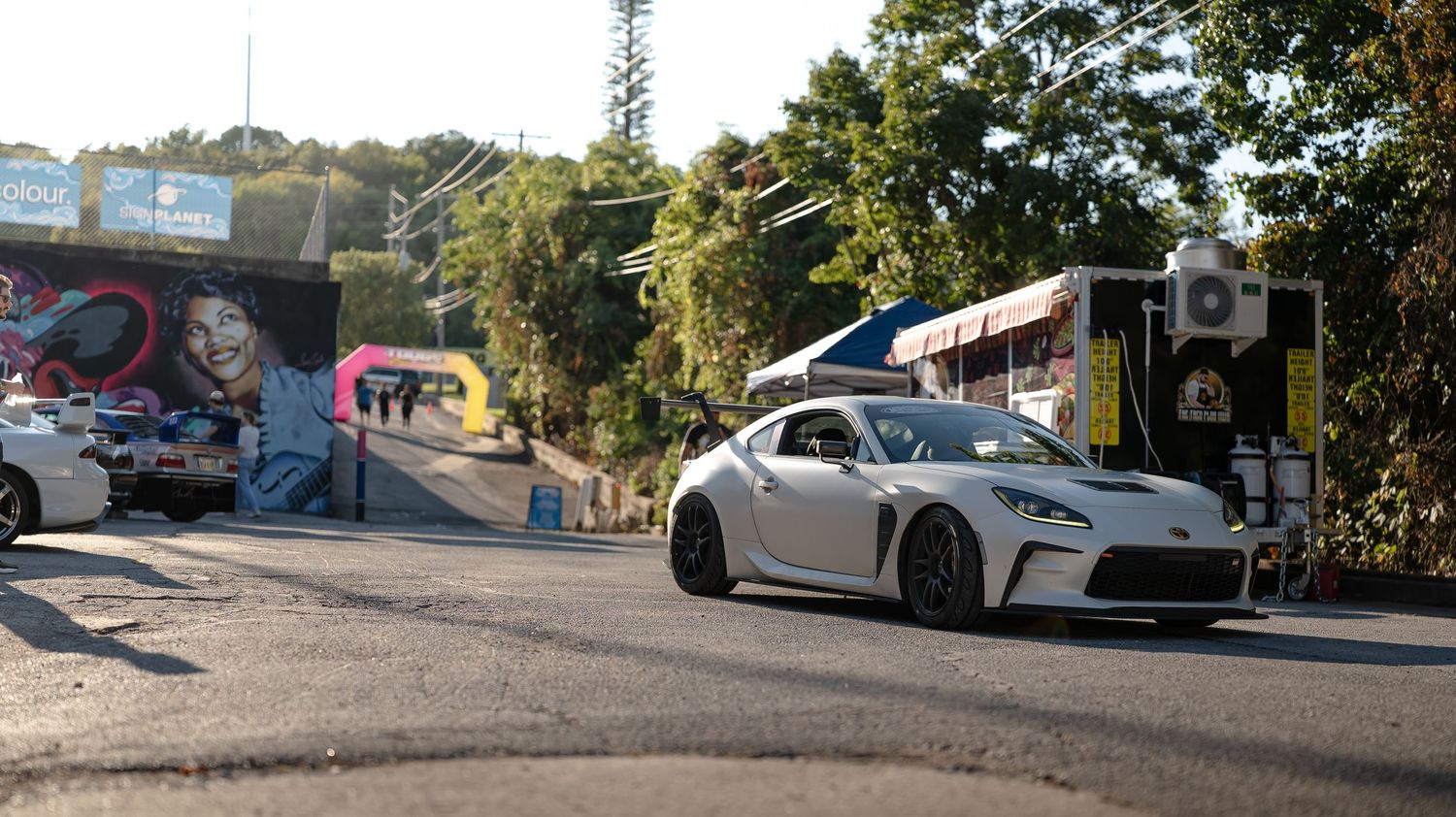 Wide panoramic view of a car meetup location with vehicles and urban scenery.