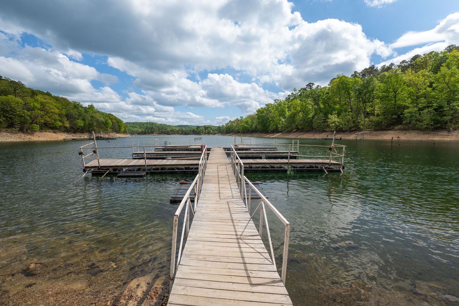 Wooden dock extending into a calm lake surrounded by lush green trees under a partly cloudy blue sky.