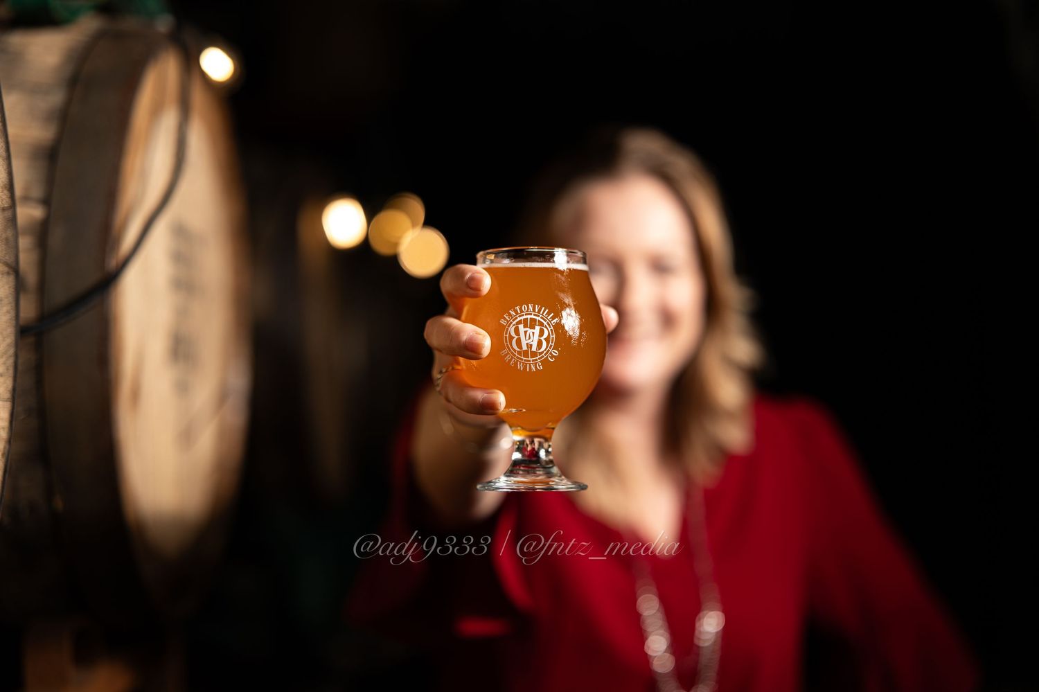 Woman in red dress holding up a craft beer glass in a dimly lit brewery with wooden barrels in background.