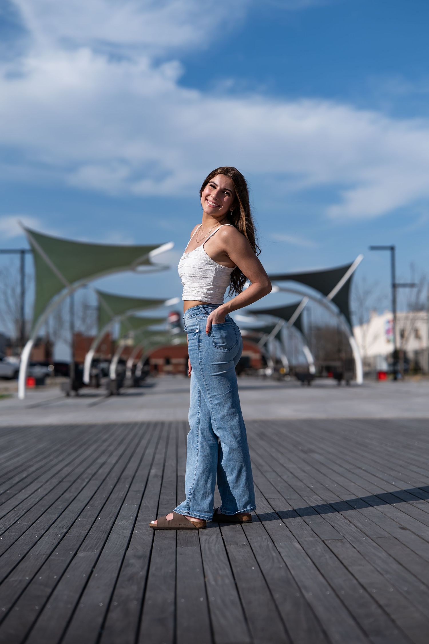 Smiling young woman in white crop top and flared jeans standing on wooden boardwalk with green canopy structures behind her.