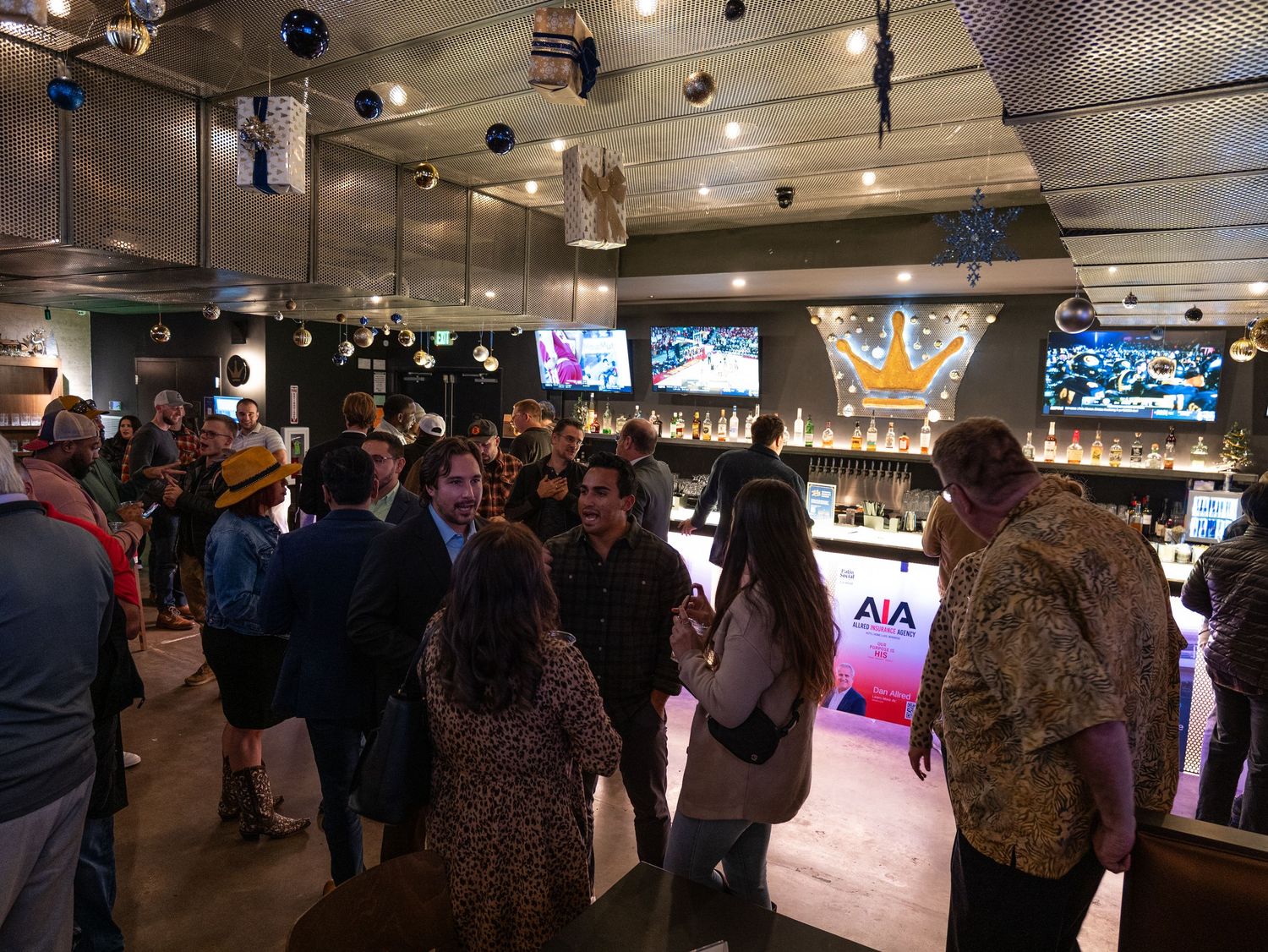 Crowded sports bar interior with multiple TV screens and patrons gathered around watching games in dim lighting.