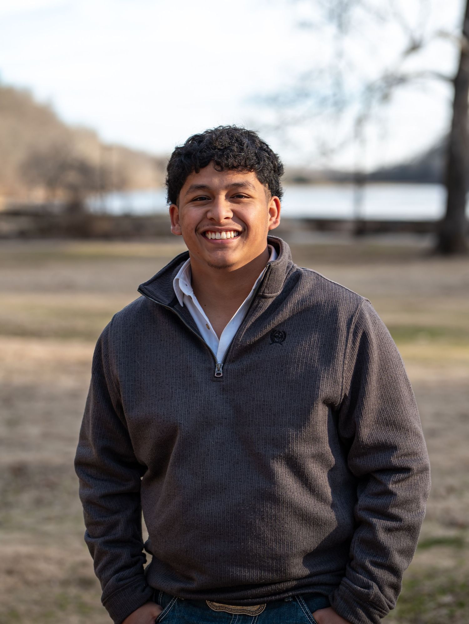 A person in a dark gray sweater stands outdoors in a field with a warm smile against a blurred natural background.