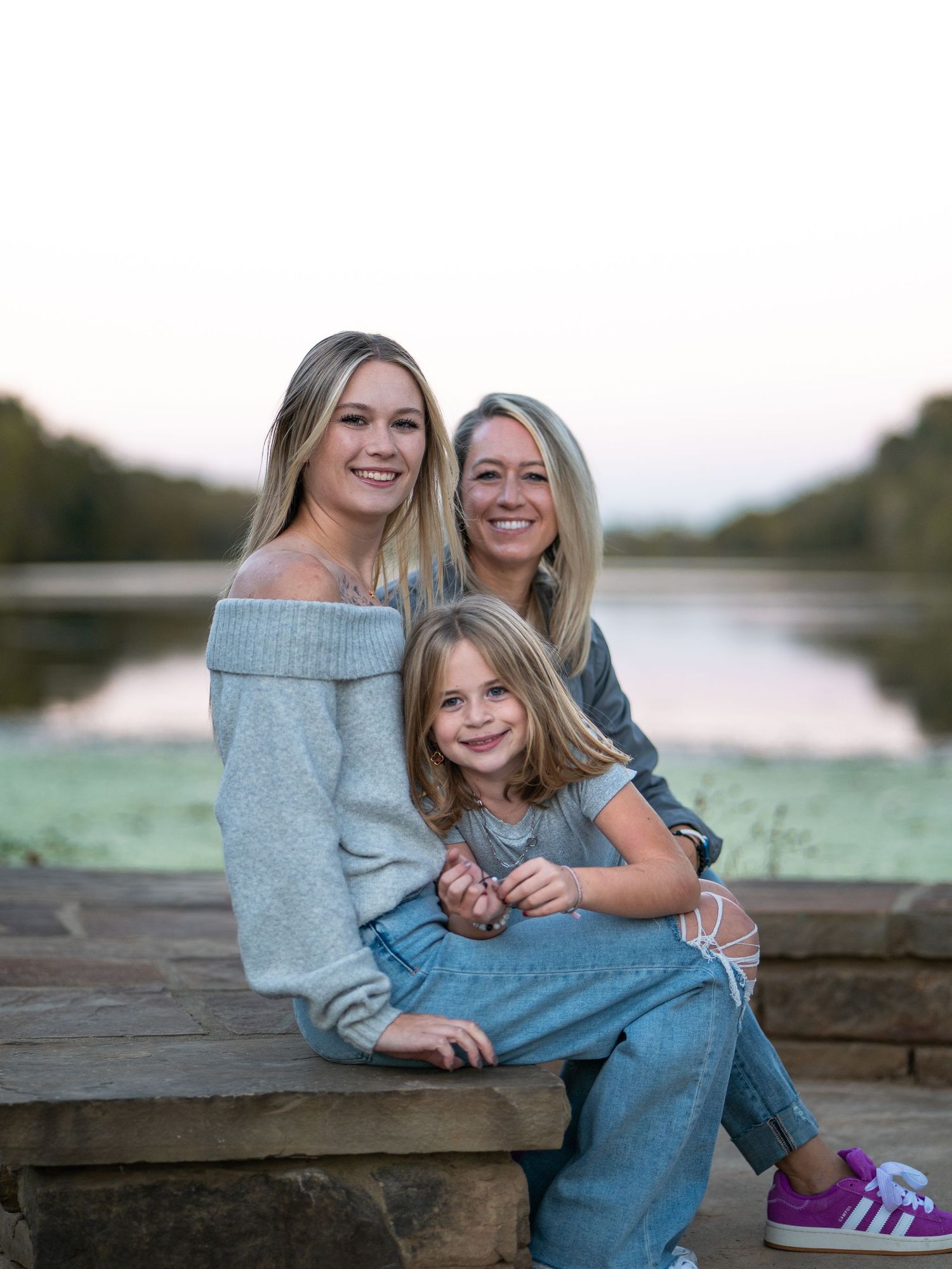 Three people sitting together on a wooden dock by a serene lake at sunset wearing casual denim and sweaters.