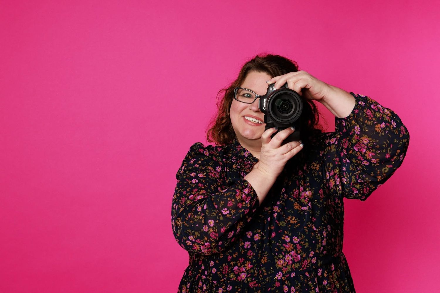 Photographer holding camera against bright pink background in floral dress.
