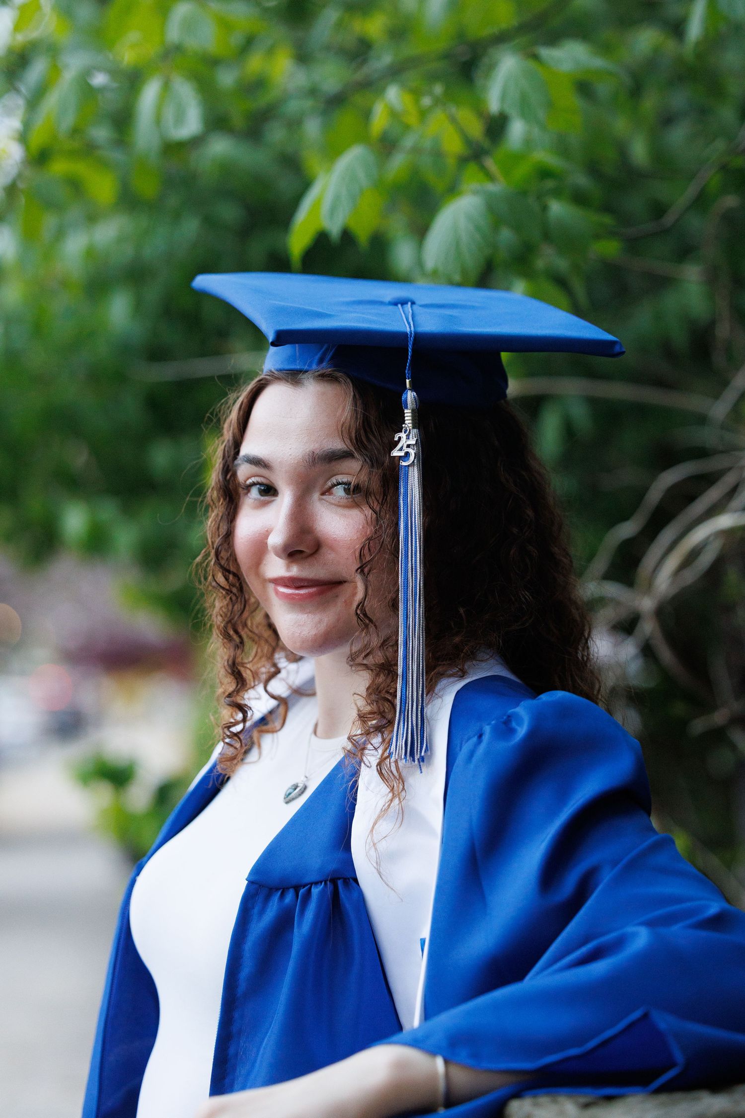 Graduate posing in blue academic regalia against green foliage background.