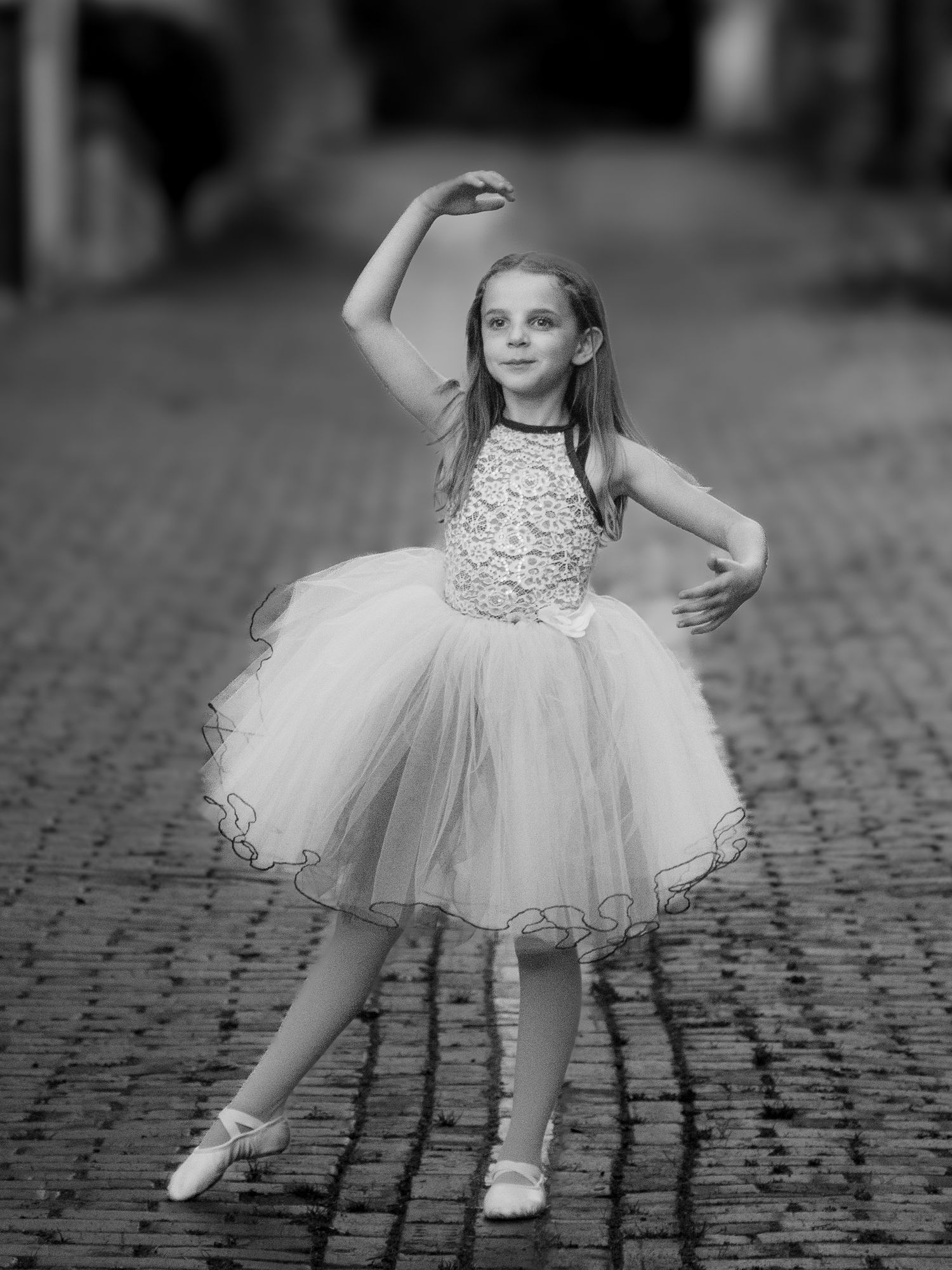 Black and white portrait of young dancer in tulle dress striking elegant pose on cobblestone street.