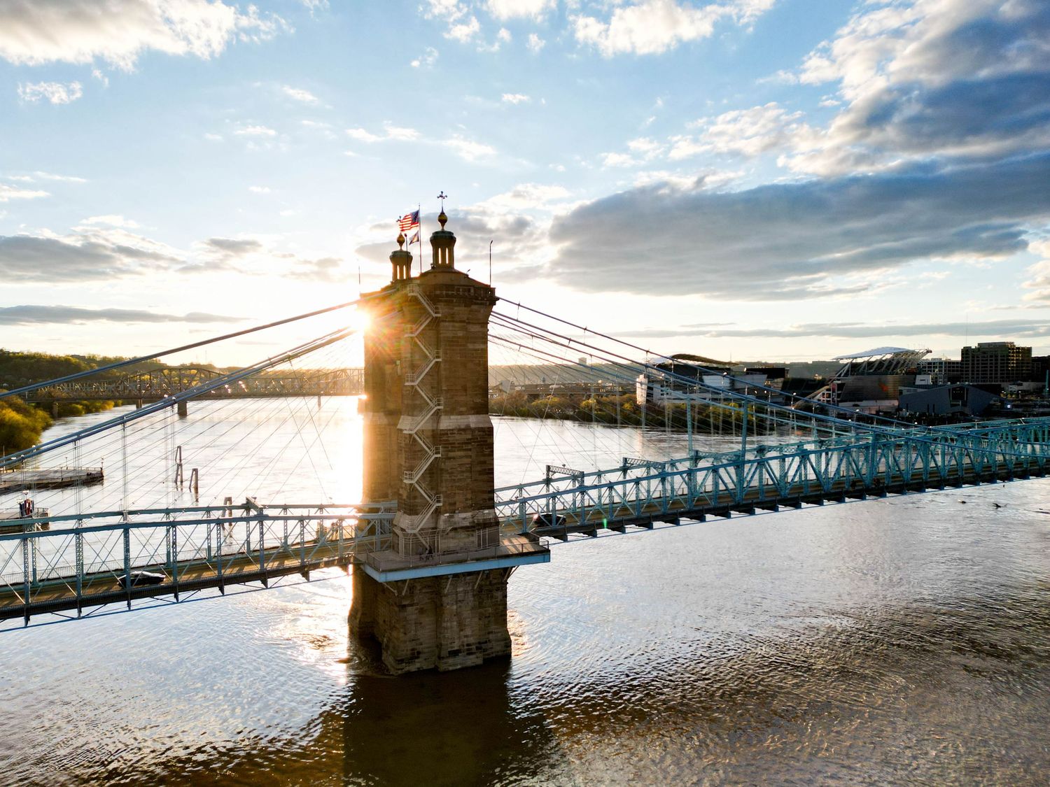 Suspension bridge silhouetted against dramatic sunset sky over water.