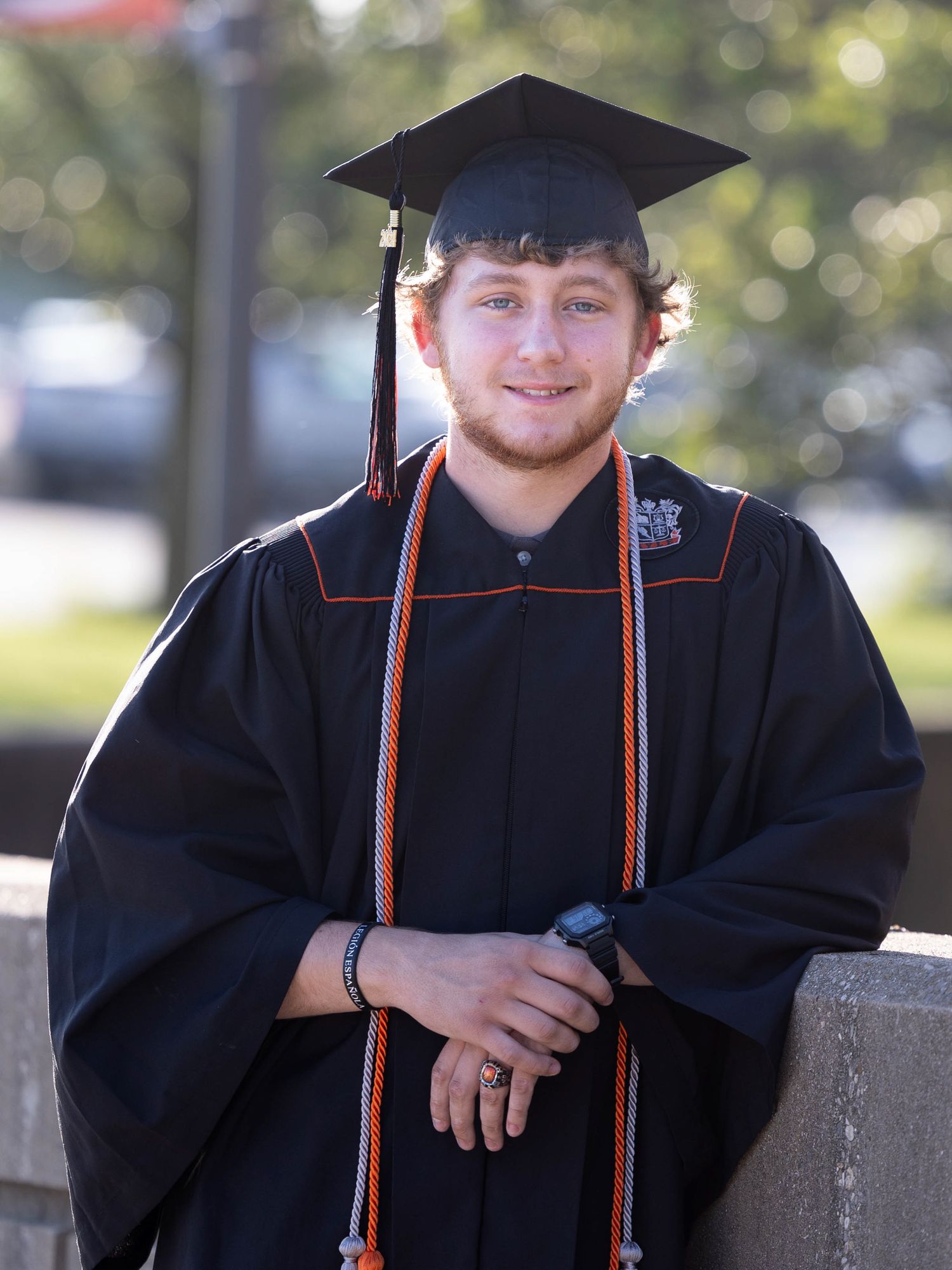 Graduate in traditional black cap and gown with orange cords outdoors.