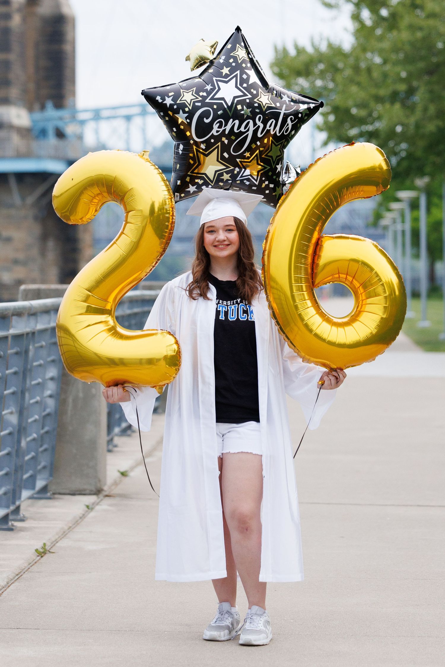 A young woman in white graduation cap and gown holds gold number 26 balloons and a Congrats star balloon outdoors.