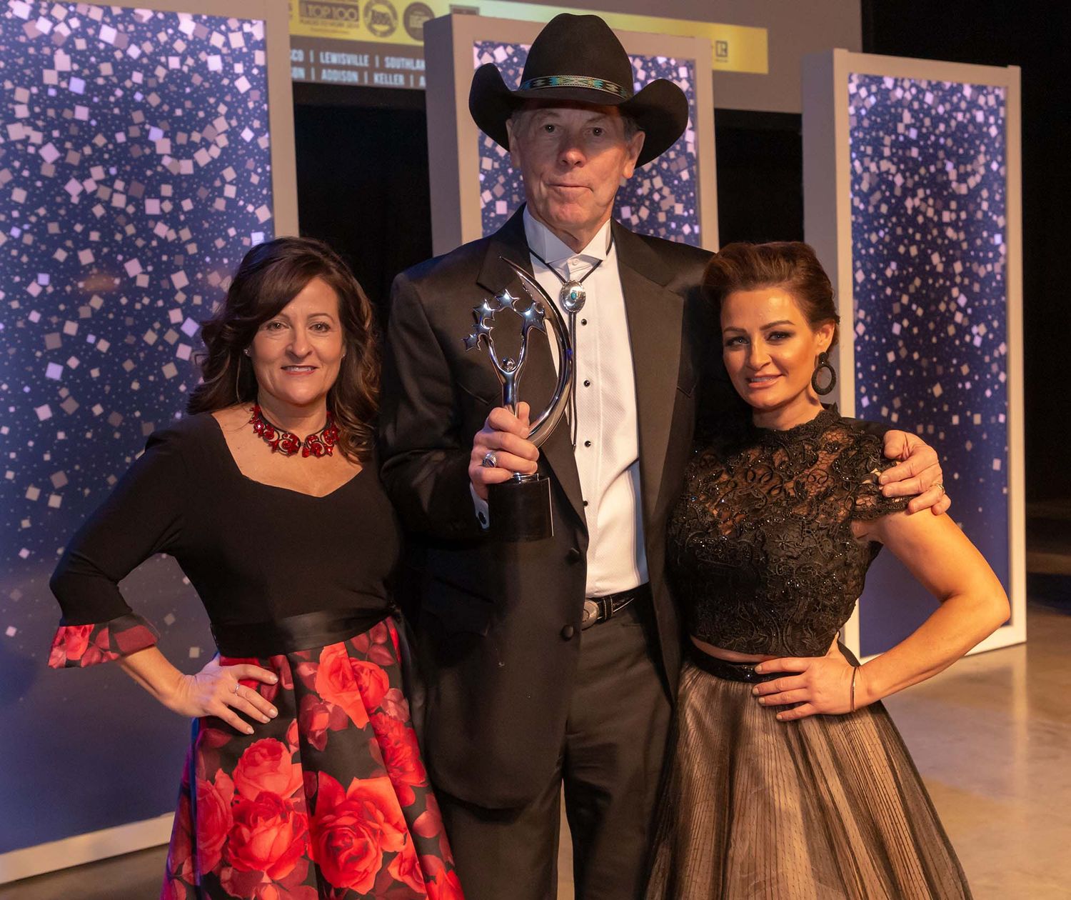 Theater performers in formal attire and red floral costumes pose on stage with sparkly backdrop.