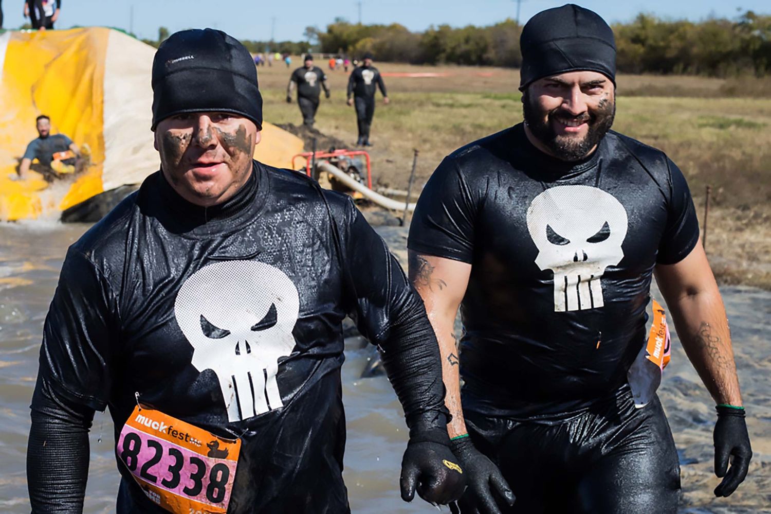 Participants wearing black athletic gear splash through muddy water during an obstacle course race.