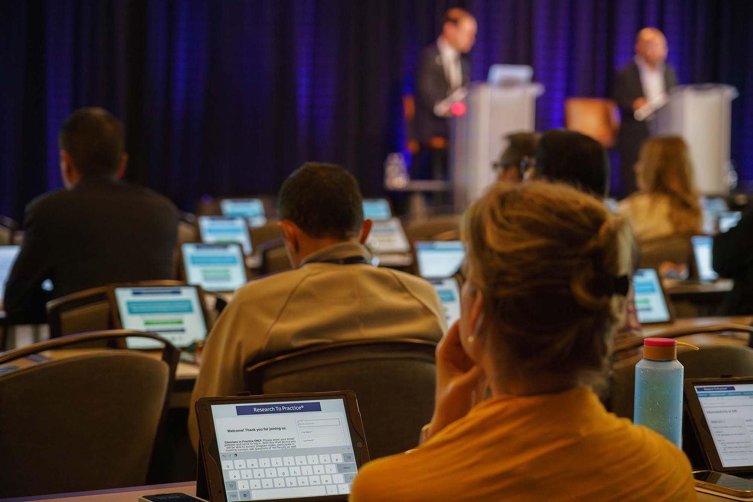 Conference attendees viewing presentation in darkened room with laptop screens visible.