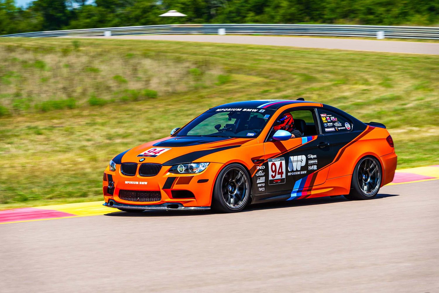 Performance racing cars including an orange BMW compete on a professional racing circuit.