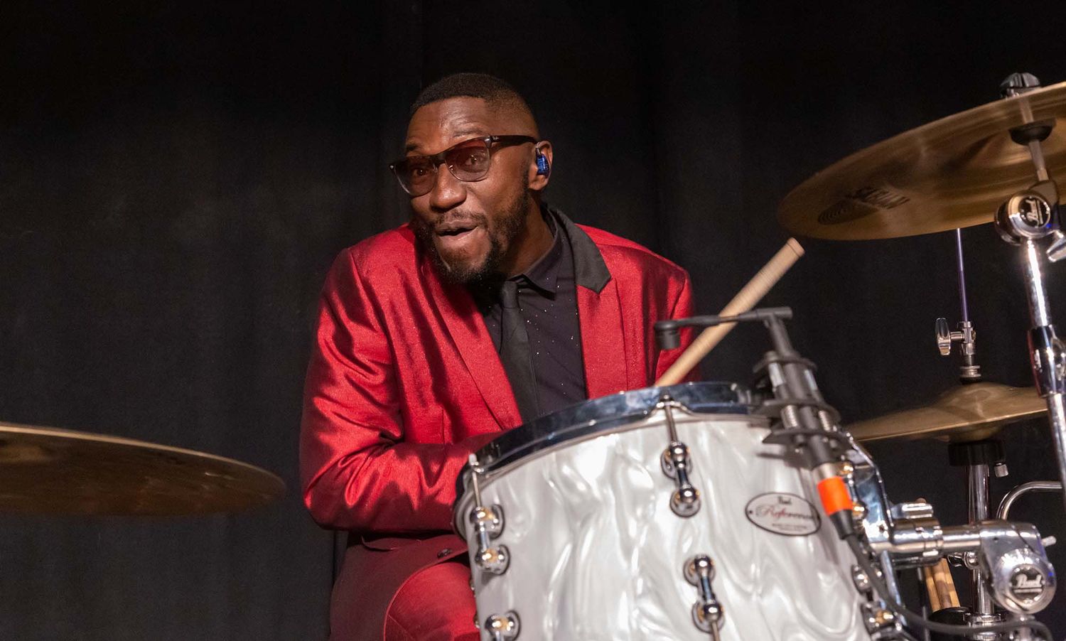 Band members in bright red outfits perform energetically on stage with drums and instruments.
