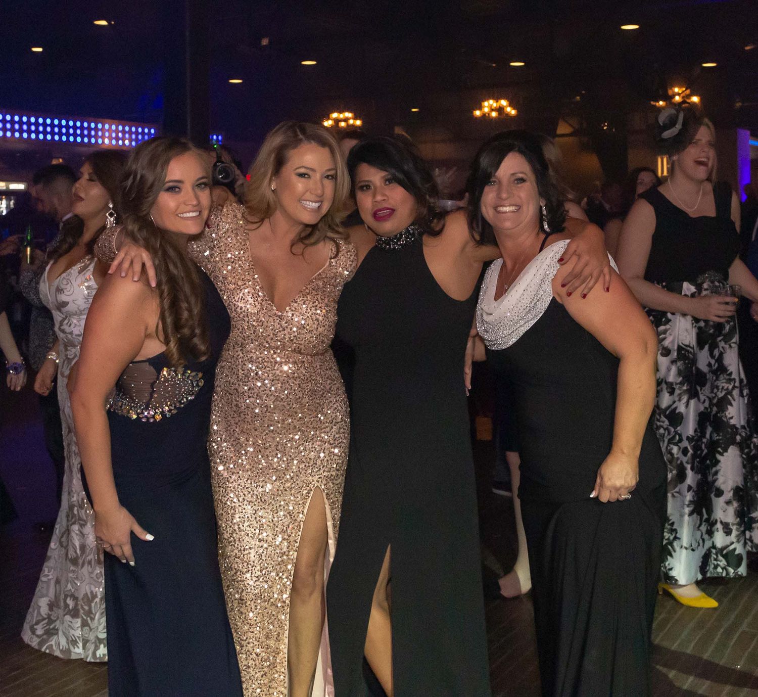 Formal event attendees mingle and pose together at an evening celebration with dark ambient lighting.