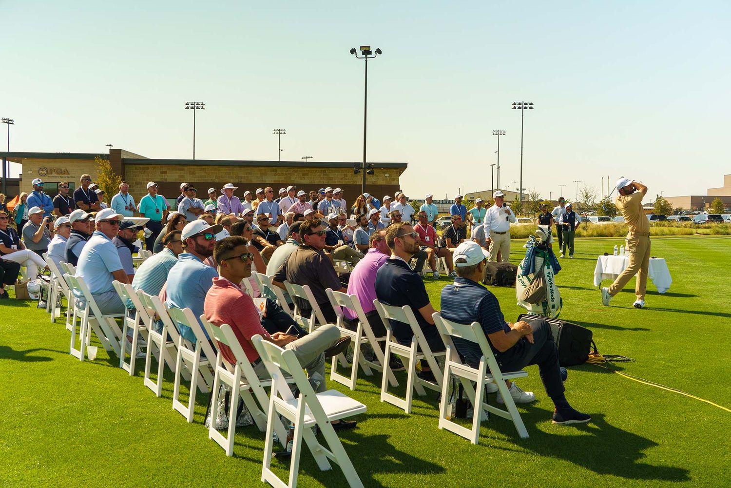 Wide view of an outdoor event space with white chairs and audience on a grass field.