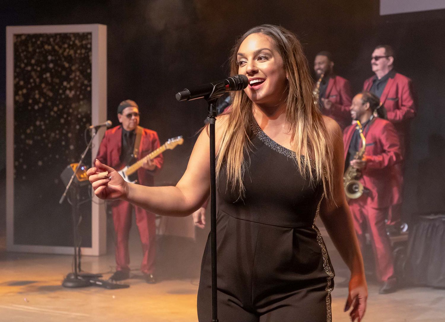 Performer in black dress sings on stage with band members in red suits in background.