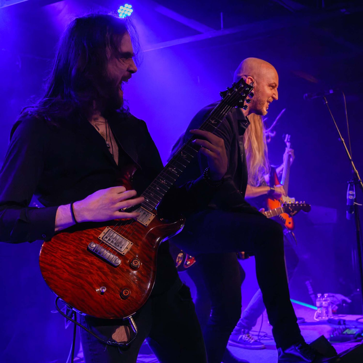Guitar players perform under purple stage lighting during rock concert.