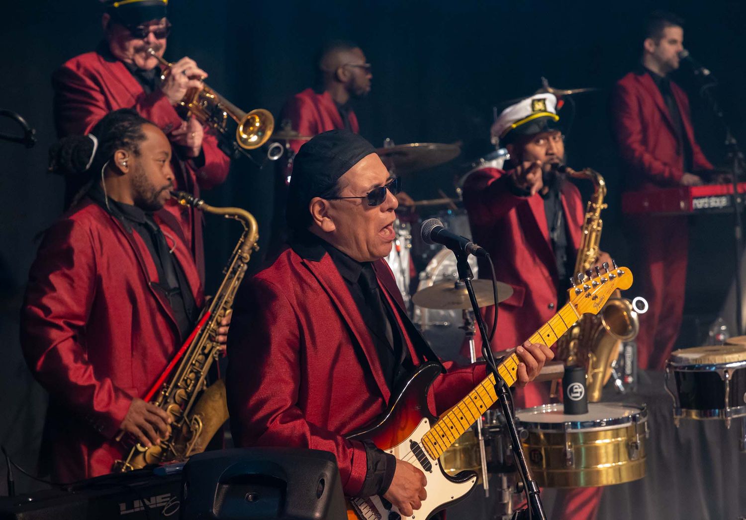 Musicians in red uniforms performing with saxophones and guitars on a dark stage.