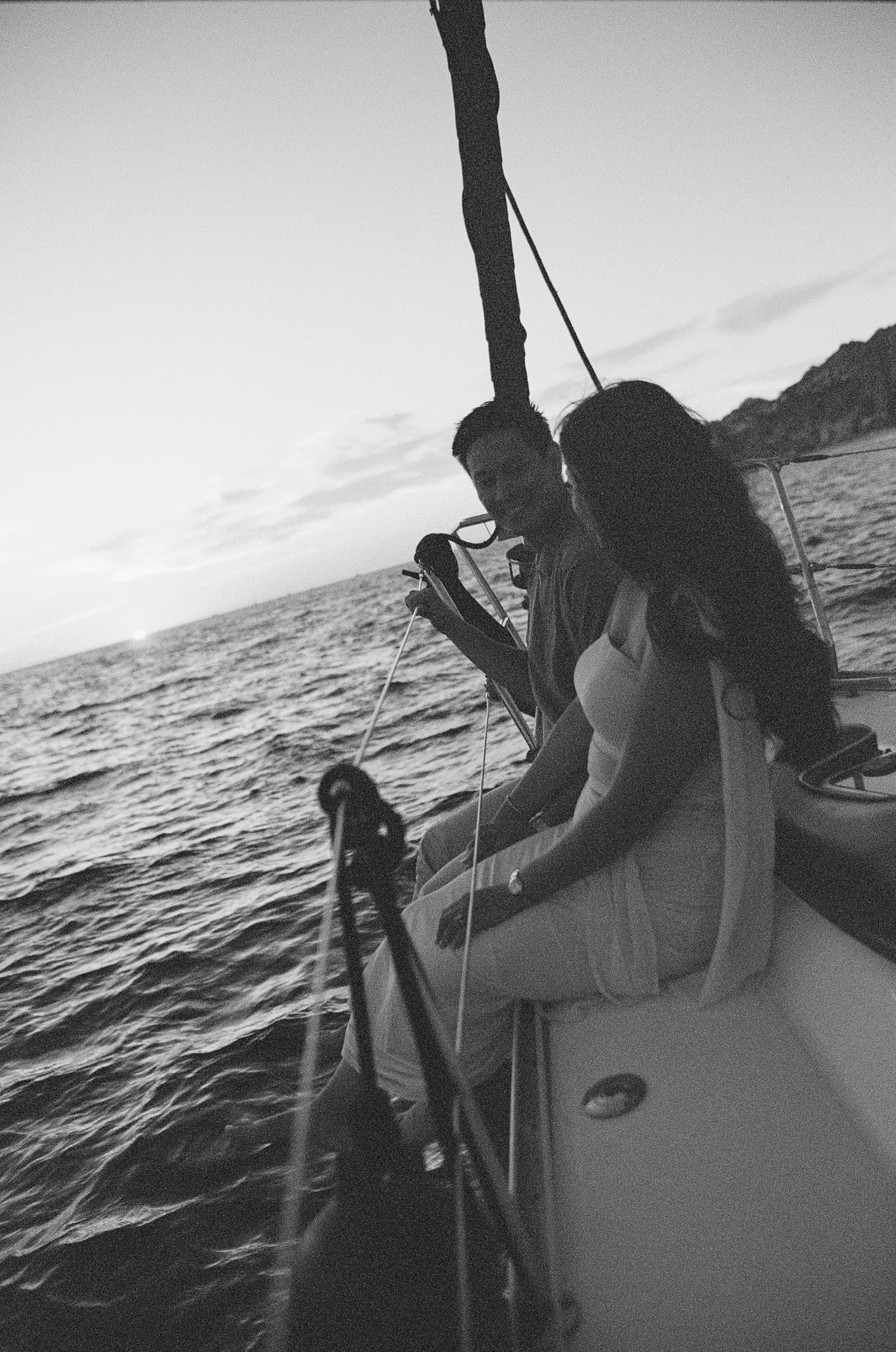 Black and white photo of a couple sitting together on a sailboat at dusk with calm ocean waters in the background.