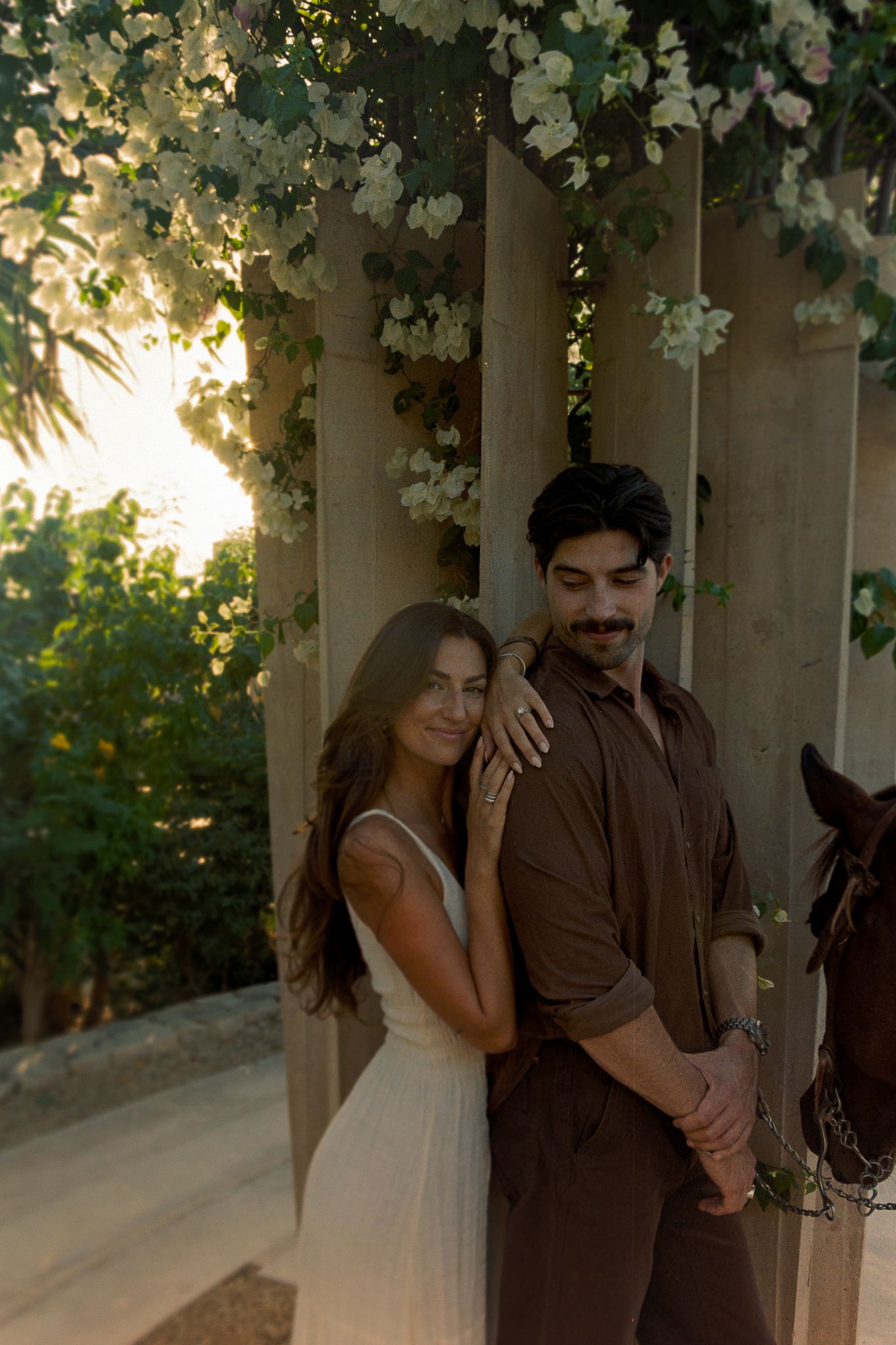 Couple embracing near floral-adorned columns at golden hour, woman in white dress, man in brown shirt, lush greenery backdrop at Acre Resort