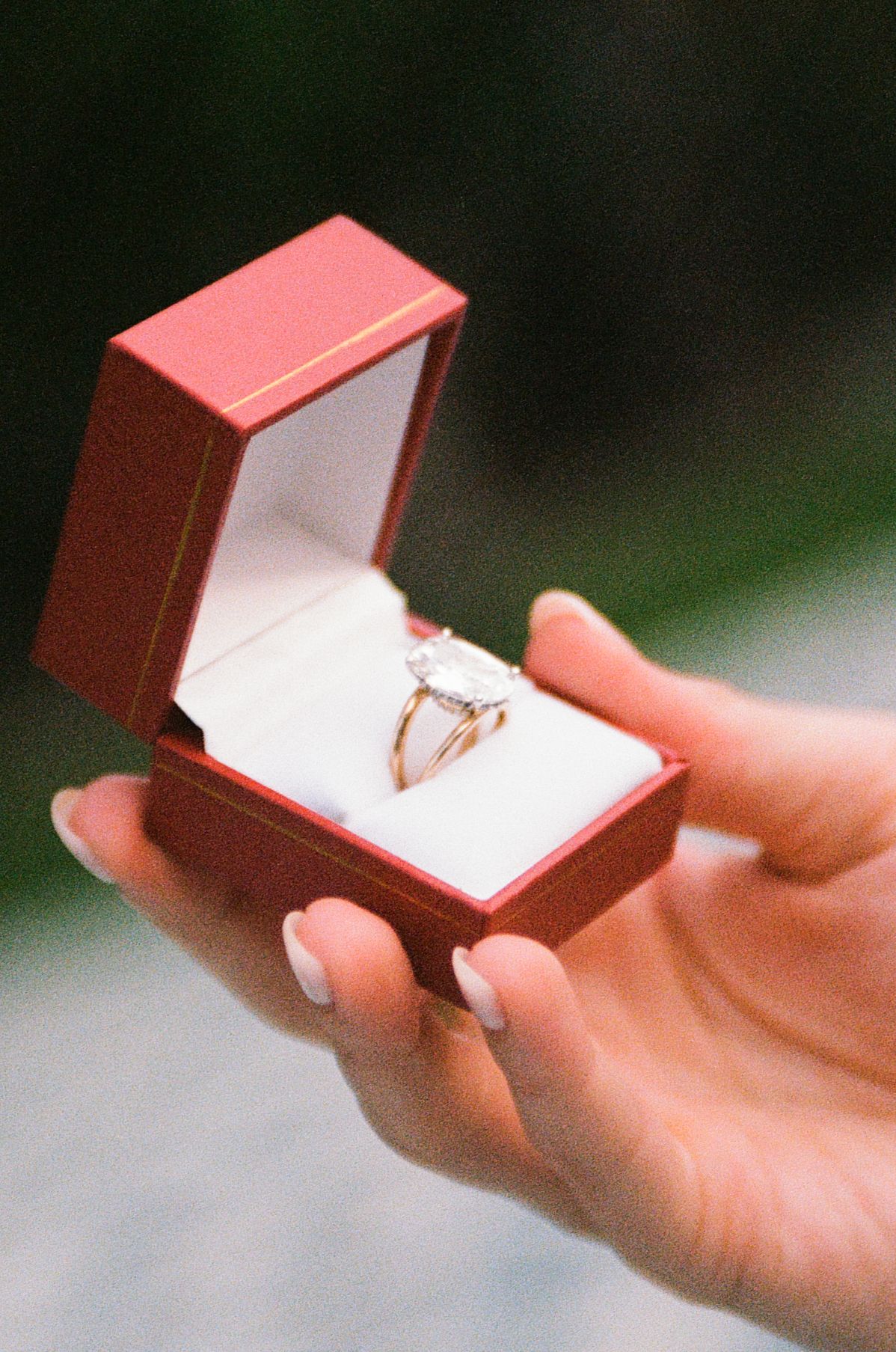 A hand holds an open red jewelry box displaying a diamond solitaire engagement ring on a white cushion.