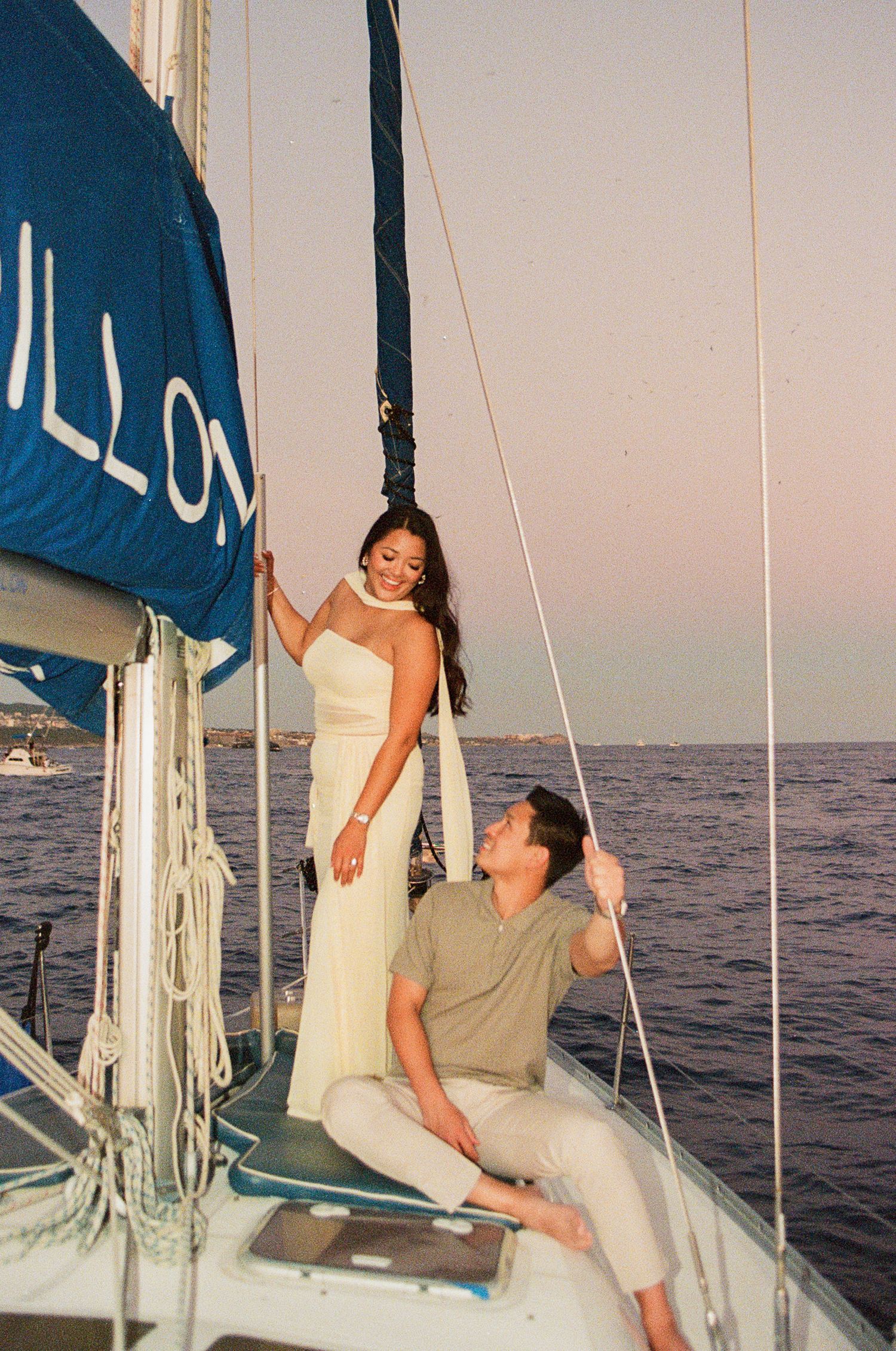 Couple posing on a sailboat at sunset, woman in white dress standing by mast, man seated looking up at her in Cabo San Lucas.