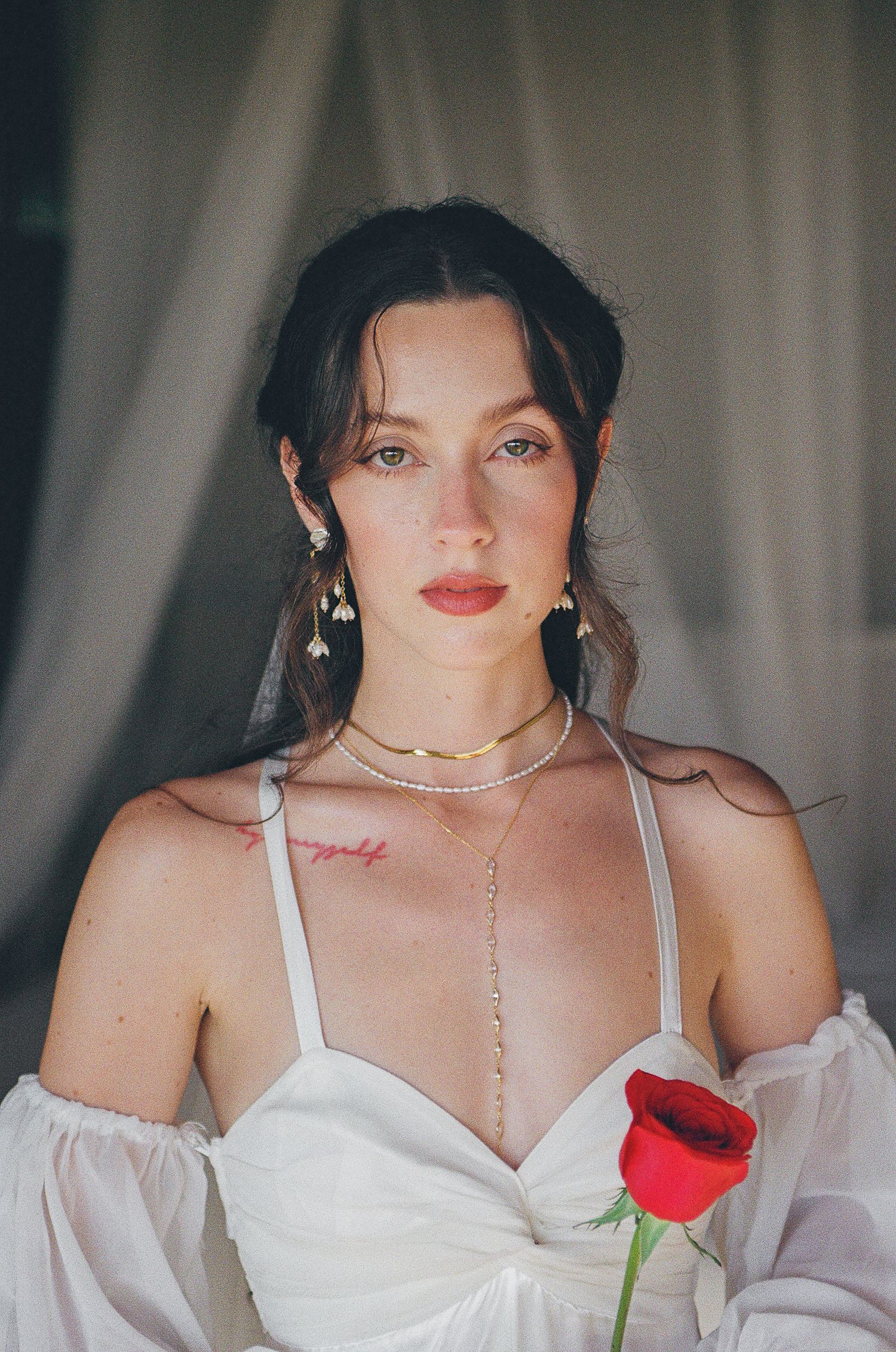 Young bride in white dress holding red rose, wearing gold jewelry and floral earrings getting ready for her elopement in Todos Santos.