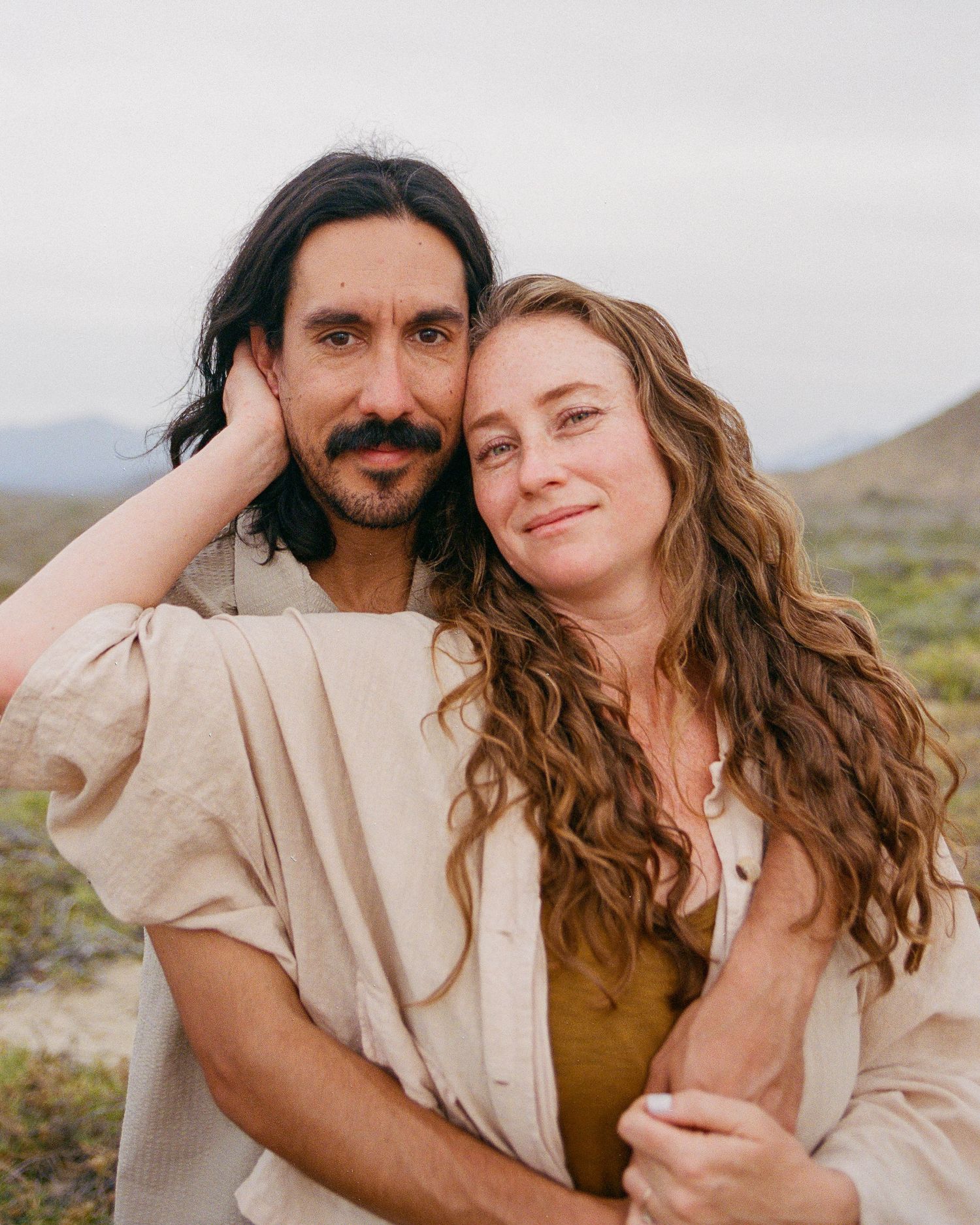 A couple embraces outdoors in a desert landscape, the woman leaning on the mans shoulder, both wearing neutral linen clothes in Todos Santos