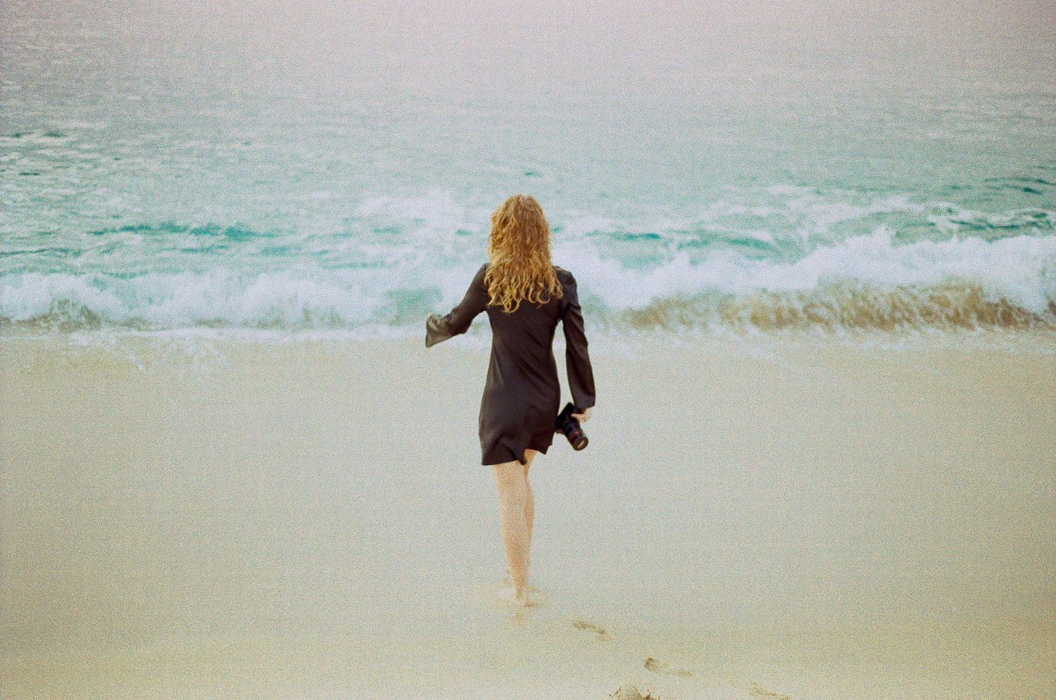 Photographer Zoey Marchiney owner of Love On Film Baja in black dress walking barefoot toward ocean waves on a sandy beach holding camera.