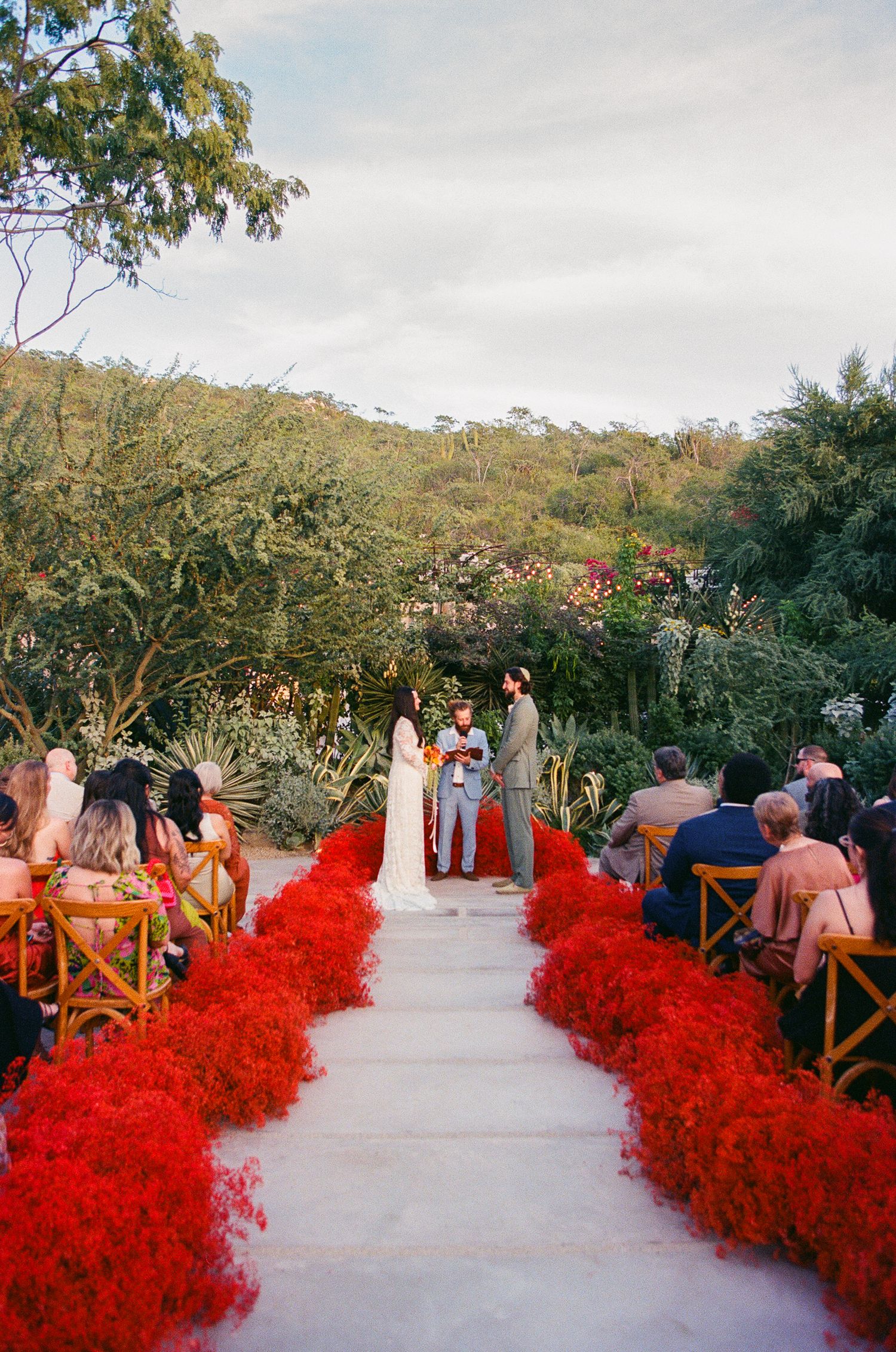 Outdoor wedding ceremony with red flower-lined aisle, couple at altar, lush greenery backdrop, and guests seated in wooden chairs at Acre.