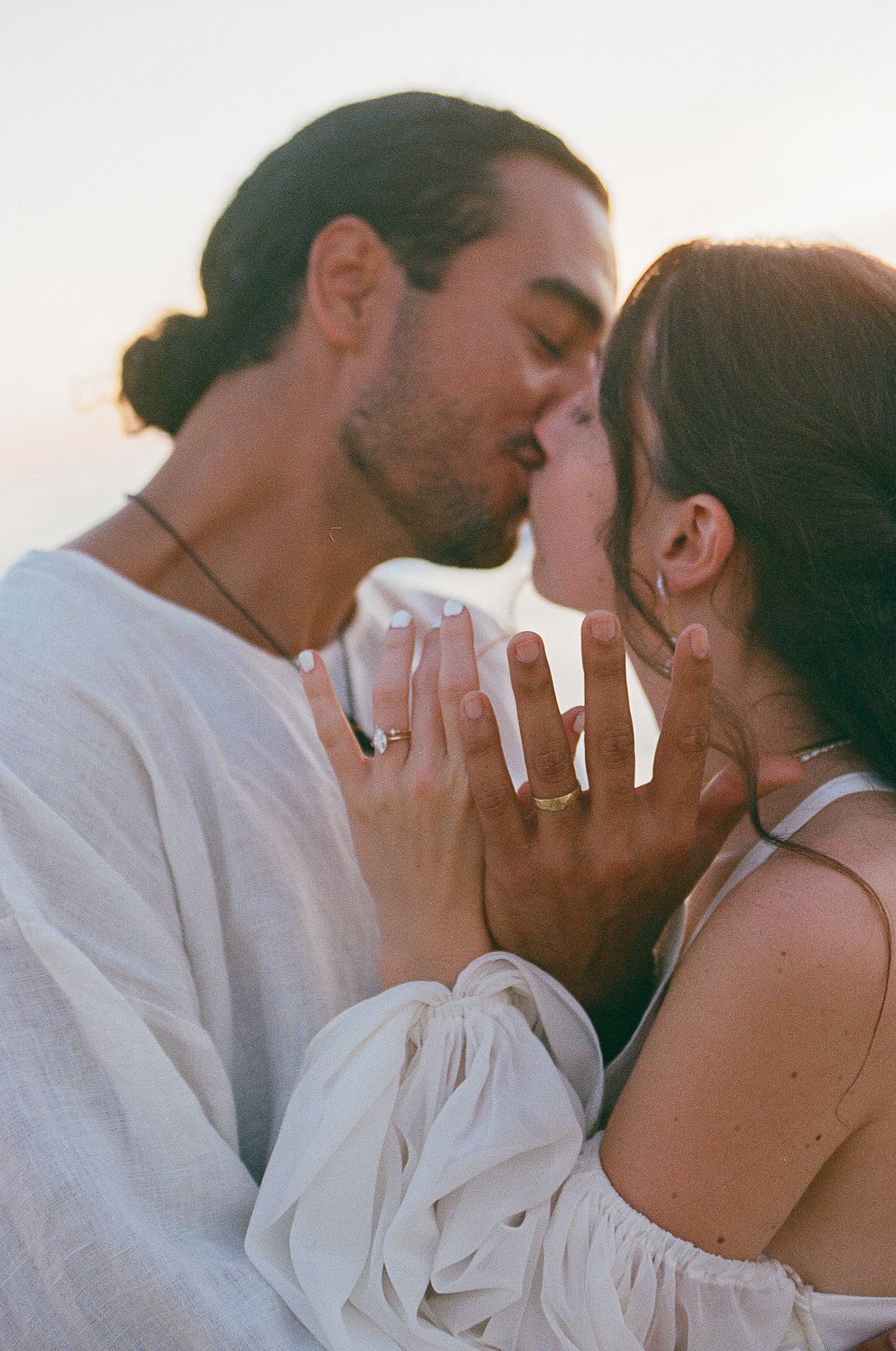 Couple sharing a romantic kiss at sunset, showing off engagement ring and wedding band on intertwined hands.